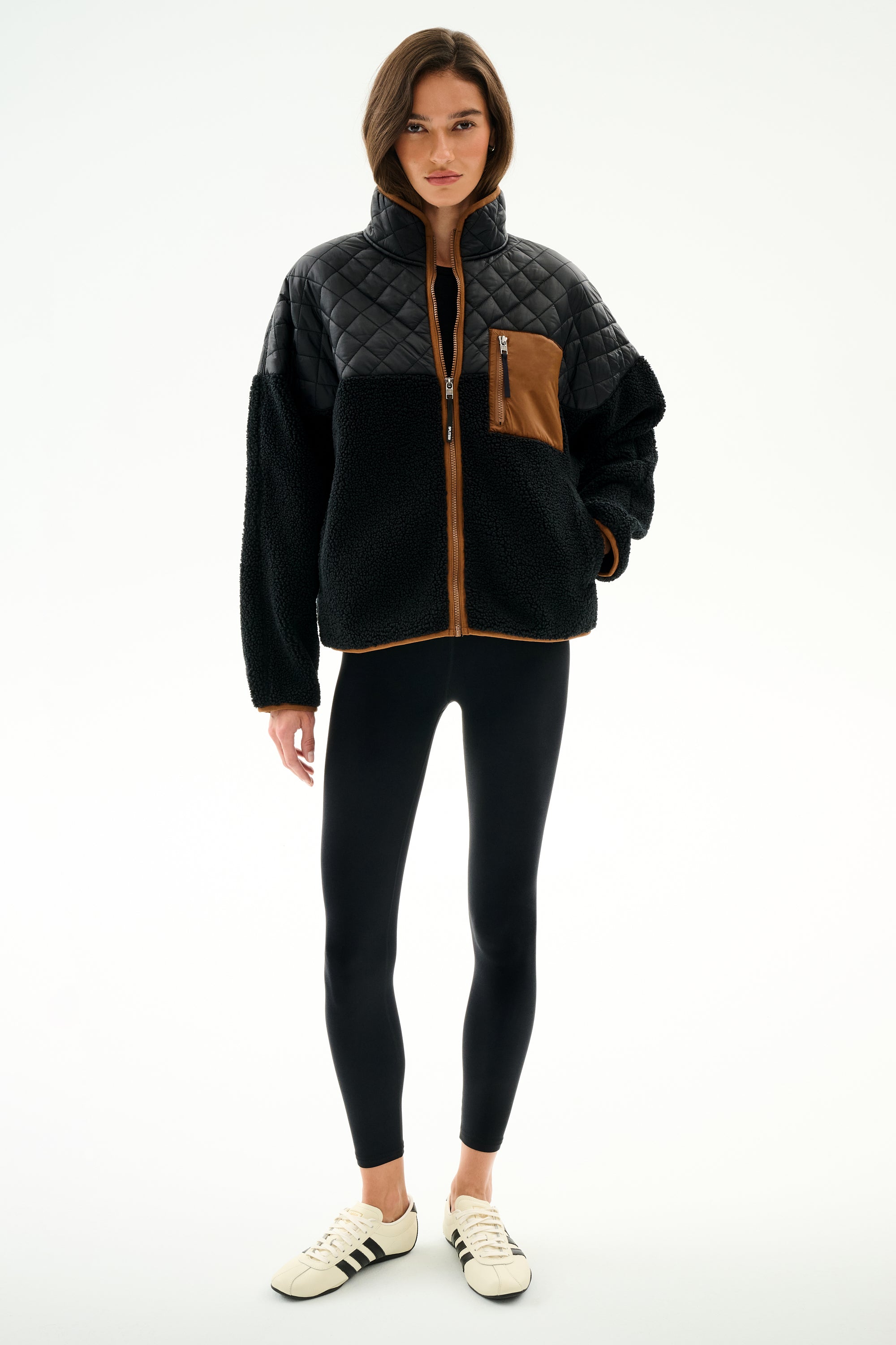 A woman wears the SPLITS59 Skylar Sherpa Jacket - Black/Toffee, paired with black leggings and white sneakers, standing against a white background.