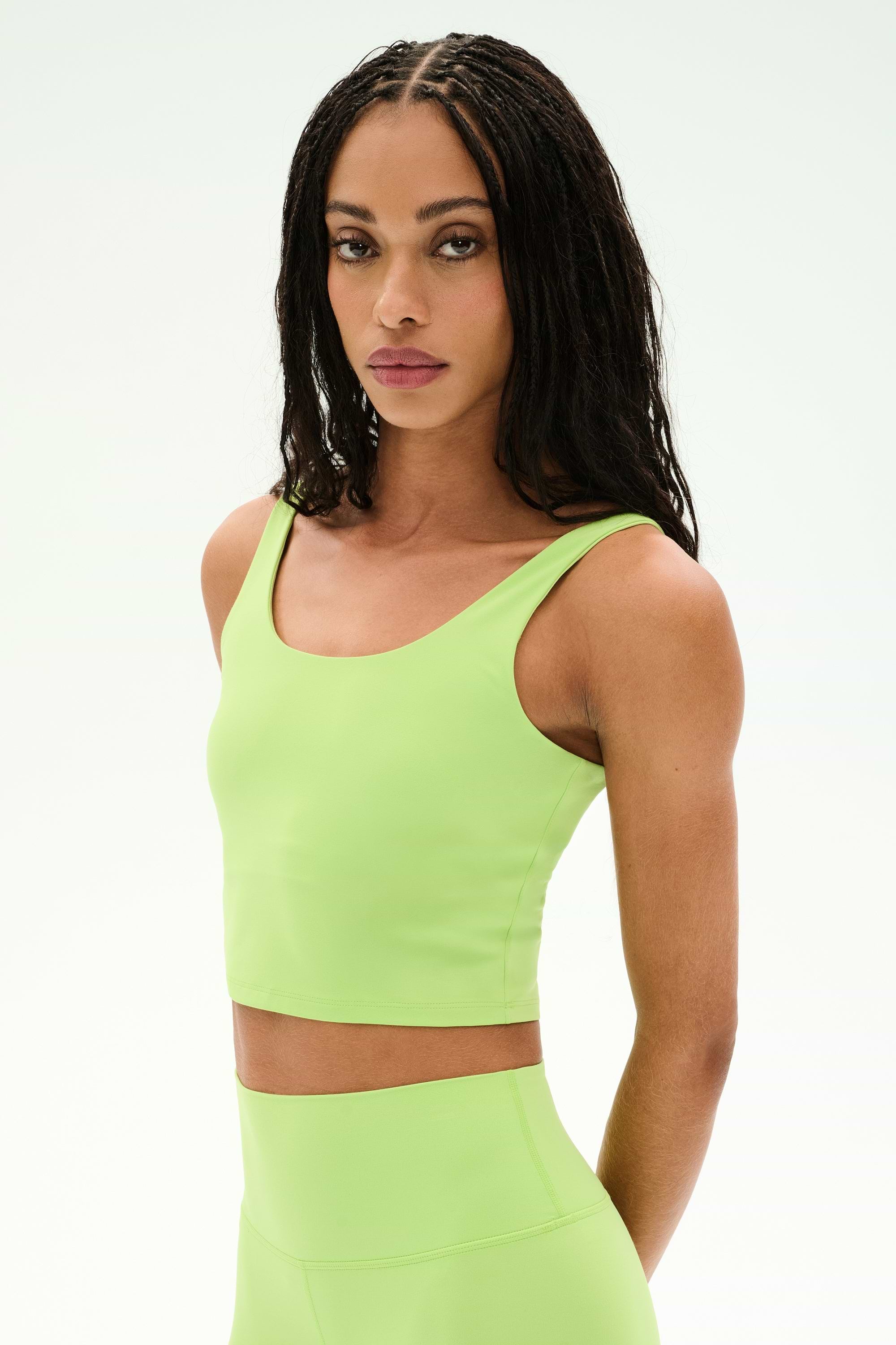 A person with long braided hair wears the SPLITS59 Sprint Rigor Bralette in Limeade—crafted from soft, stretchy nylon spandex—as they stand against a plain light background, looking at the camera.
