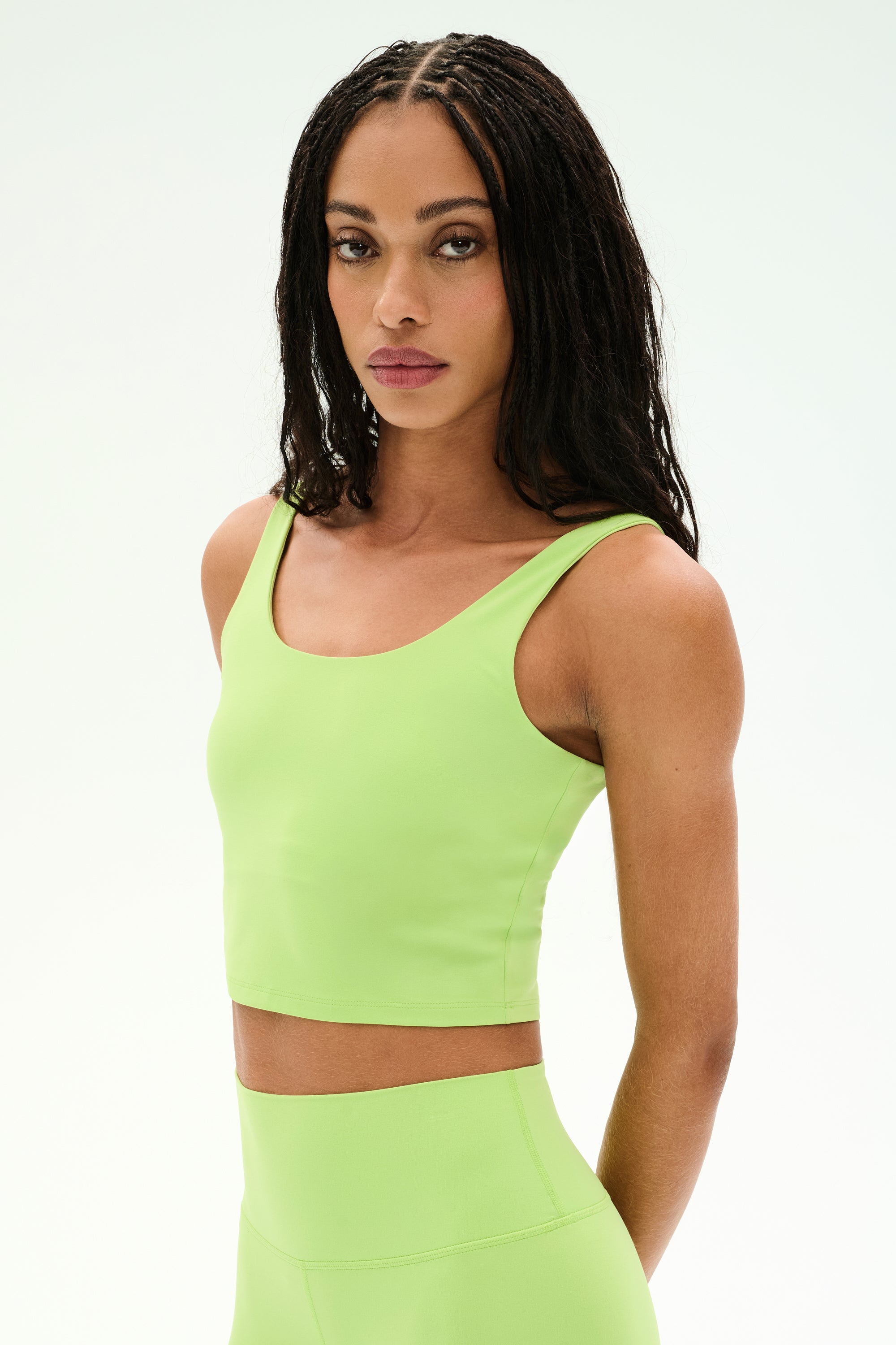 A person with long braided hair wears the SPLITS59 Sprint Rigor Bralette in Limeade—crafted from soft, stretchy nylon spandex—as they stand against a plain light background, looking at the camera.