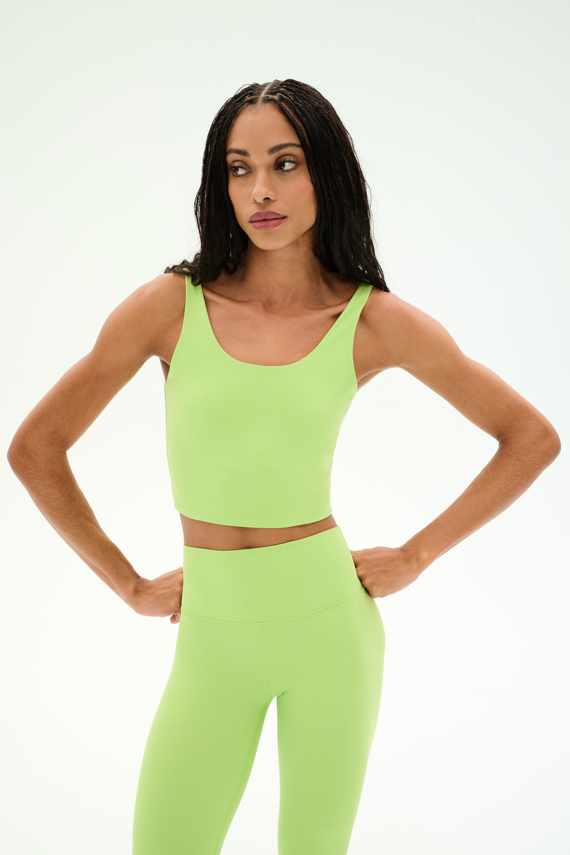 A woman stands with her hands on her hips against a plain white background, wearing the SPLITS59 Sprint Rigor Bralette in Limeade—a lime green, sleeveless crop top crafted from stretchy, figure-hugging nylon spandex fabric (62% nylon, 38% spandex).