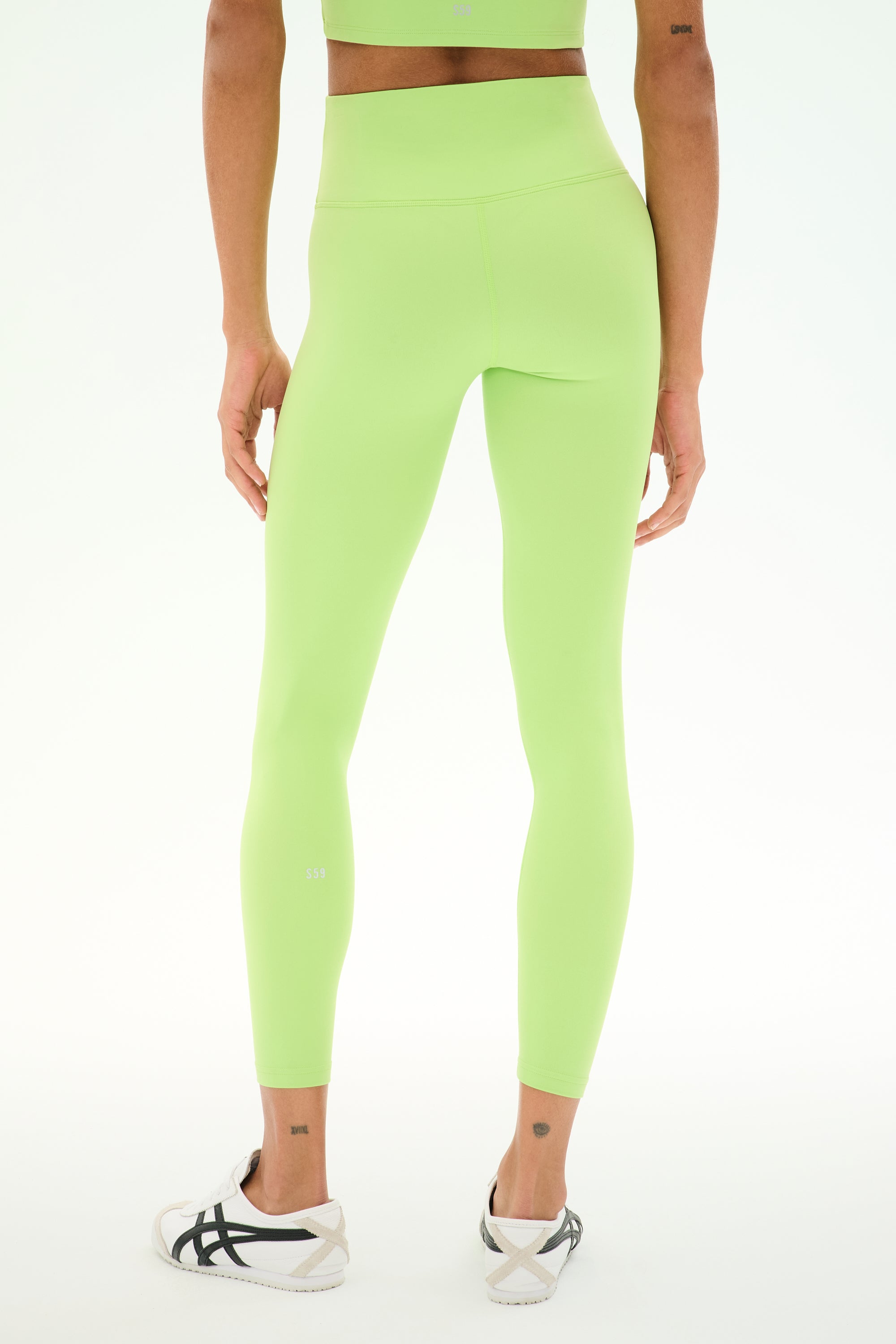 A person is seen from the back, standing against a plain background while wearing SPLITS59 Sprint High Waist Rigor 7/8 leggings in Limeade—a high-waisted, light green style crafted from stretchy nylon-spandex fabric—paired with white sneakers.