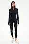 A woman stands facing forward, wearing the SPLITS59 Tess Rigor Half Zip Waist Length top in black, black leggings with white side stripes, and silver sneakers against a plain white background.