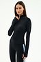 A woman with long brown hair wears the SPLITS59 Tess Rigor Half Zip Waist Length top in black, paired with matching black leggings featuring a light blue stripe, standing against a plain white background.
