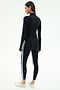 A woman faces away, wearing the SPLITS59 Tess Rigor Half Zip Waist Length top in black with matching leggings featuring white and blue side stripes, paired with white sneakers.