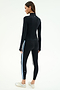 A woman faces away, wearing the SPLITS59 Tess Rigor Half Zip Waist Length top in black with matching leggings featuring white and blue side stripes, paired with white sneakers.