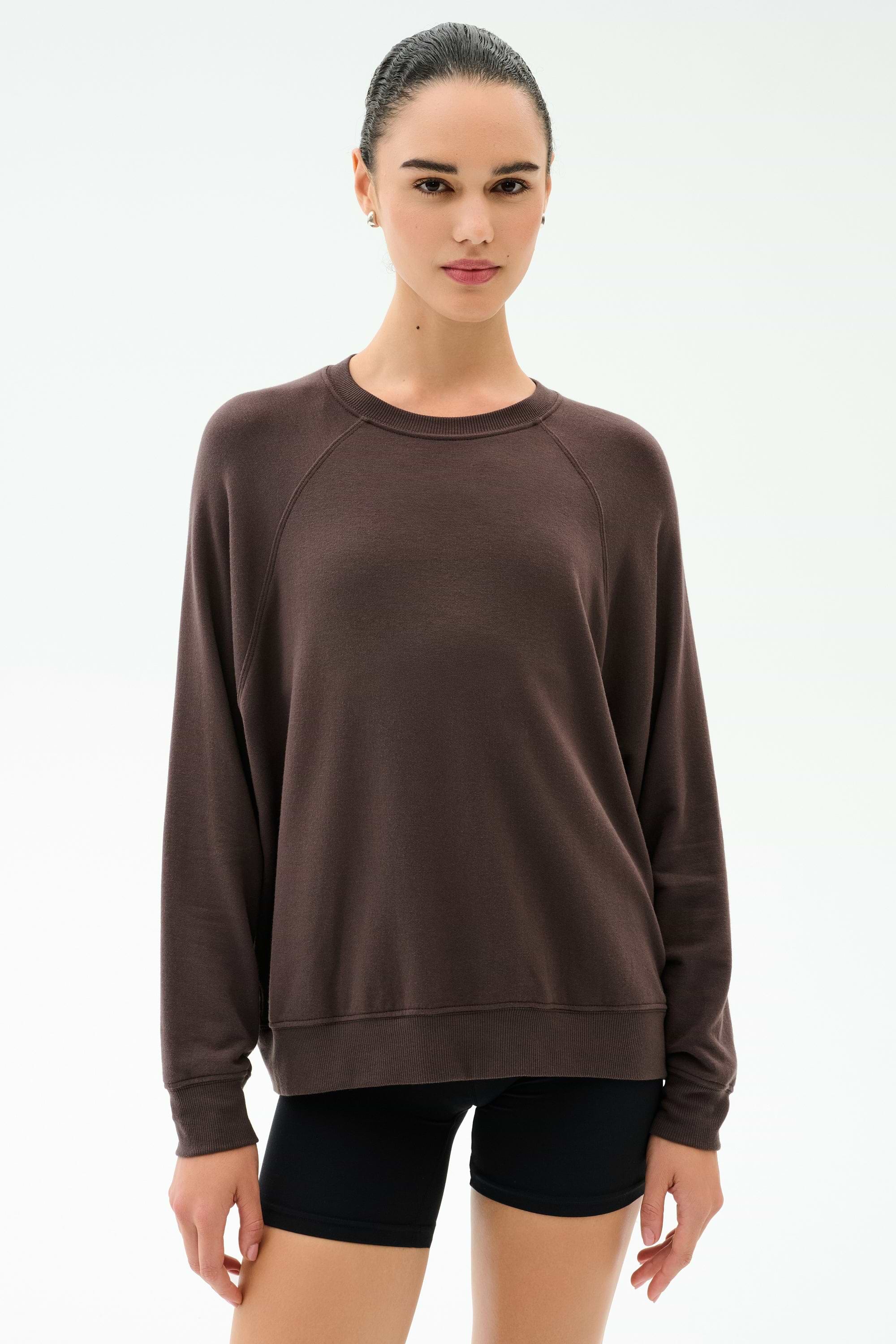 A person stands facing forward, wearing the SPLITS59 Andie Oversized Fleece Sweatshirt in Dark Chocolate and black shorts against a plain light background.