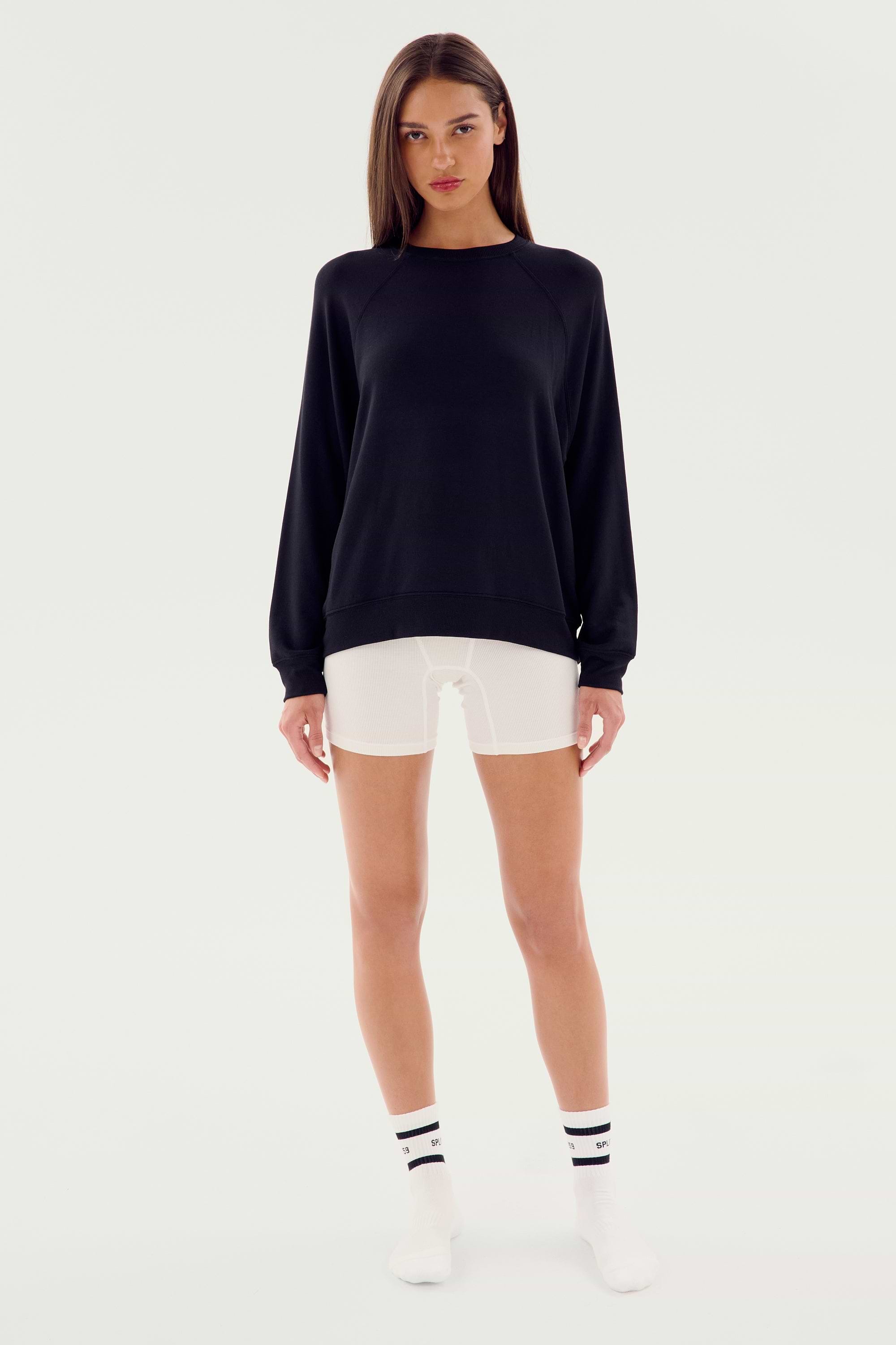 A person standing against a plain background, wearing an SPLITS59 Andie Fleece Sweatshirt - Black made in Los Angeles from stretch modal fabric, white shorts, white socks with black stripes, and white shoes.