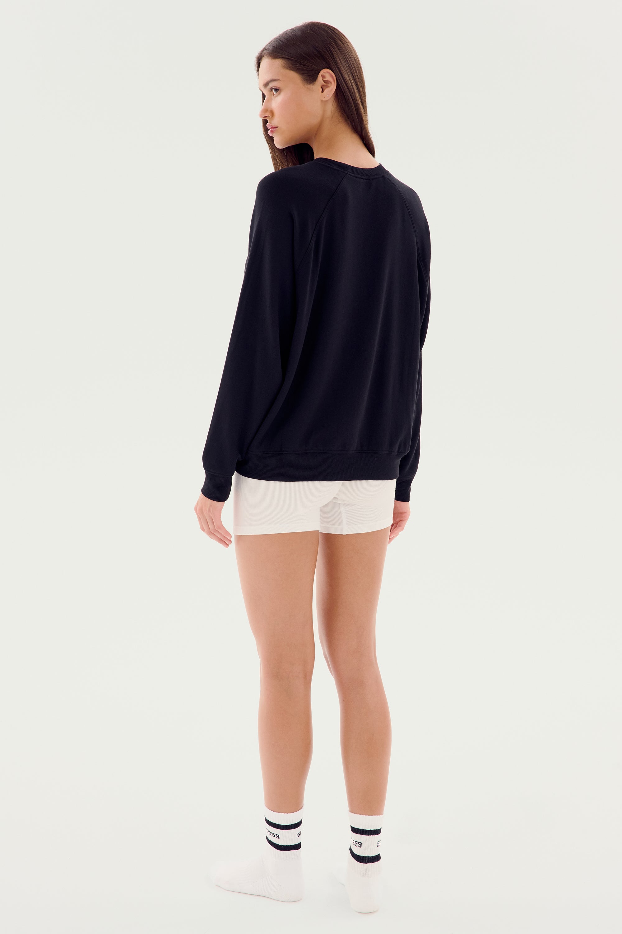 A person stands facing away, wearing a SPLITS59 Andie Fleece Sweatshirt - Black, white shorts, and white socks against a plain background.