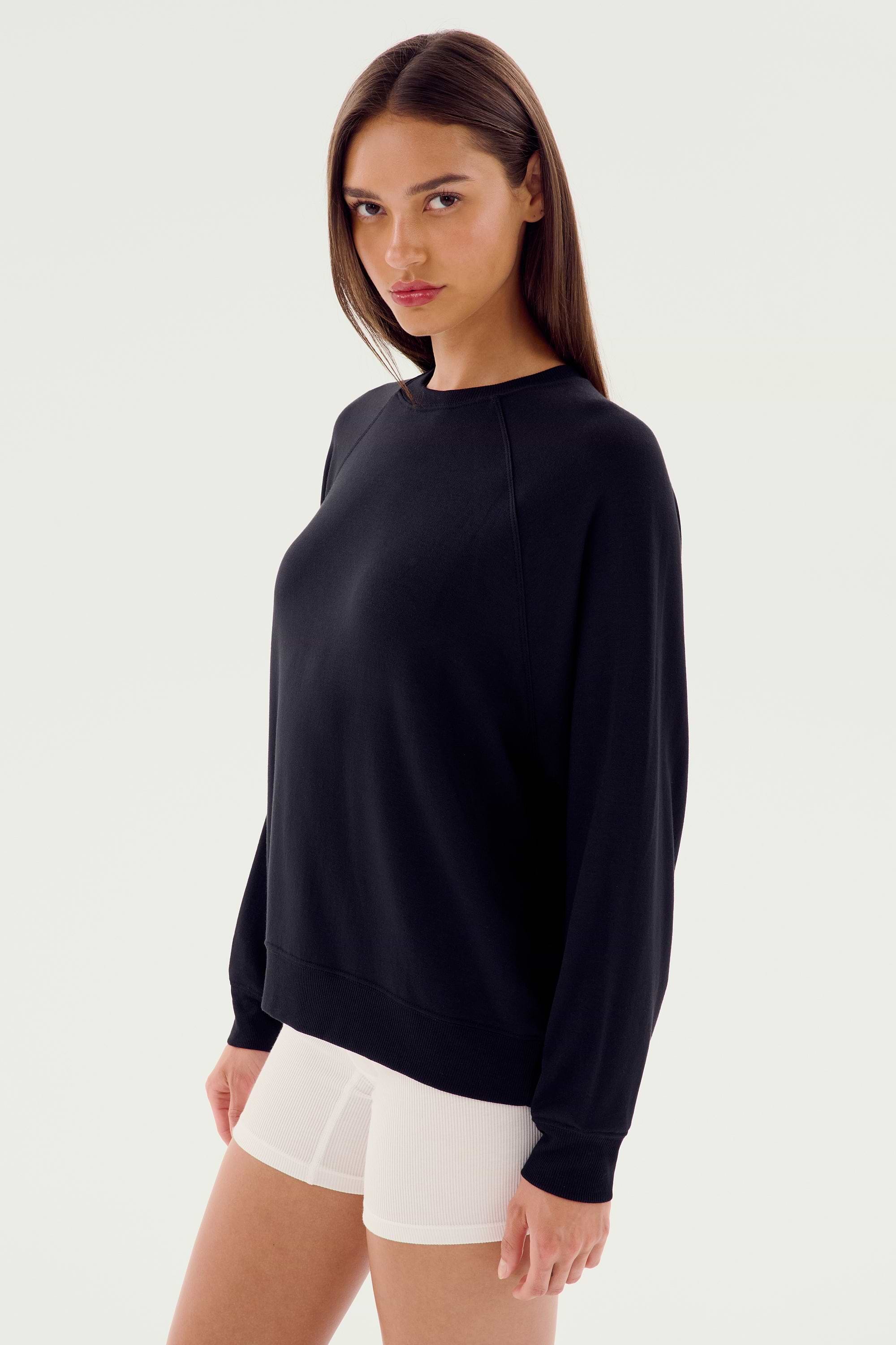 A person stands wearing the SPLITS59 Andie Fleece Sweatshirt - Black and white shorts, facing the camera with a neutral expression. The background is plain and white.