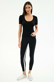 A woman stands against a plain background, wearing the SPLITS59 Ashby Rib Short Sleeve in black, black leggings with a side stripe, and white sneakers.