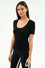 Woman with straight brown hair wearing the SPLITS59 Ashby Rib Short Sleeve top in black, paired with black leggings featuring light blue and white side stripes, standing against a plain light background.