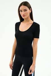 Woman with straight brown hair wearing the SPLITS59 Ashby Rib Short Sleeve top in black, paired with black leggings featuring light blue and white side stripes, standing against a plain light background.