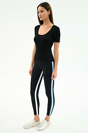 A woman stands against a white background wearing the SPLITS59 Ashby Rib Short Sleeve in black, paired with black leggings featuring white and blue side stripes and white sneakers.
