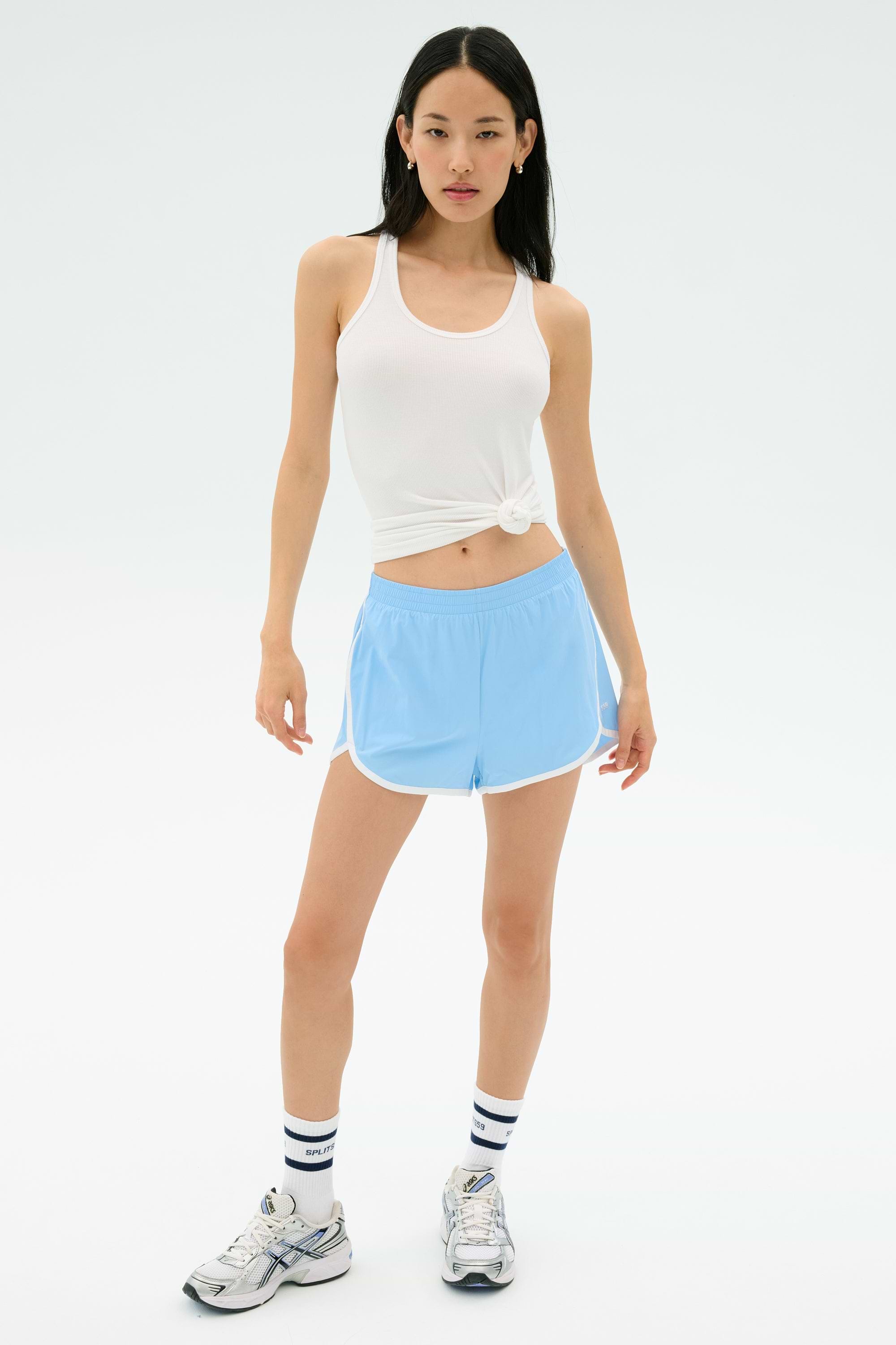 A woman wears the Splits59 Ashby Rib Tank in white, paired with light blue athletic shorts, striped socks, and sneakers, posing against a plain white background.