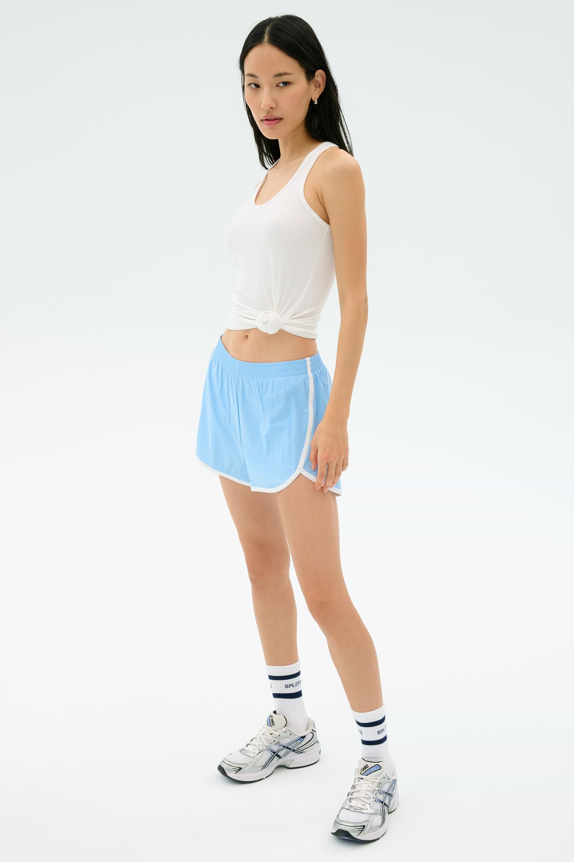 A woman wears the Splits59 Ashby Rib Tank in white, paired with light blue athletic shorts, striped white socks, and running shoes, standing against a plain light background.
