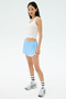 A woman wears the Splits59 Ashby Rib Tank in white, paired with light blue athletic shorts, striped white socks, and running shoes, standing against a plain light background.