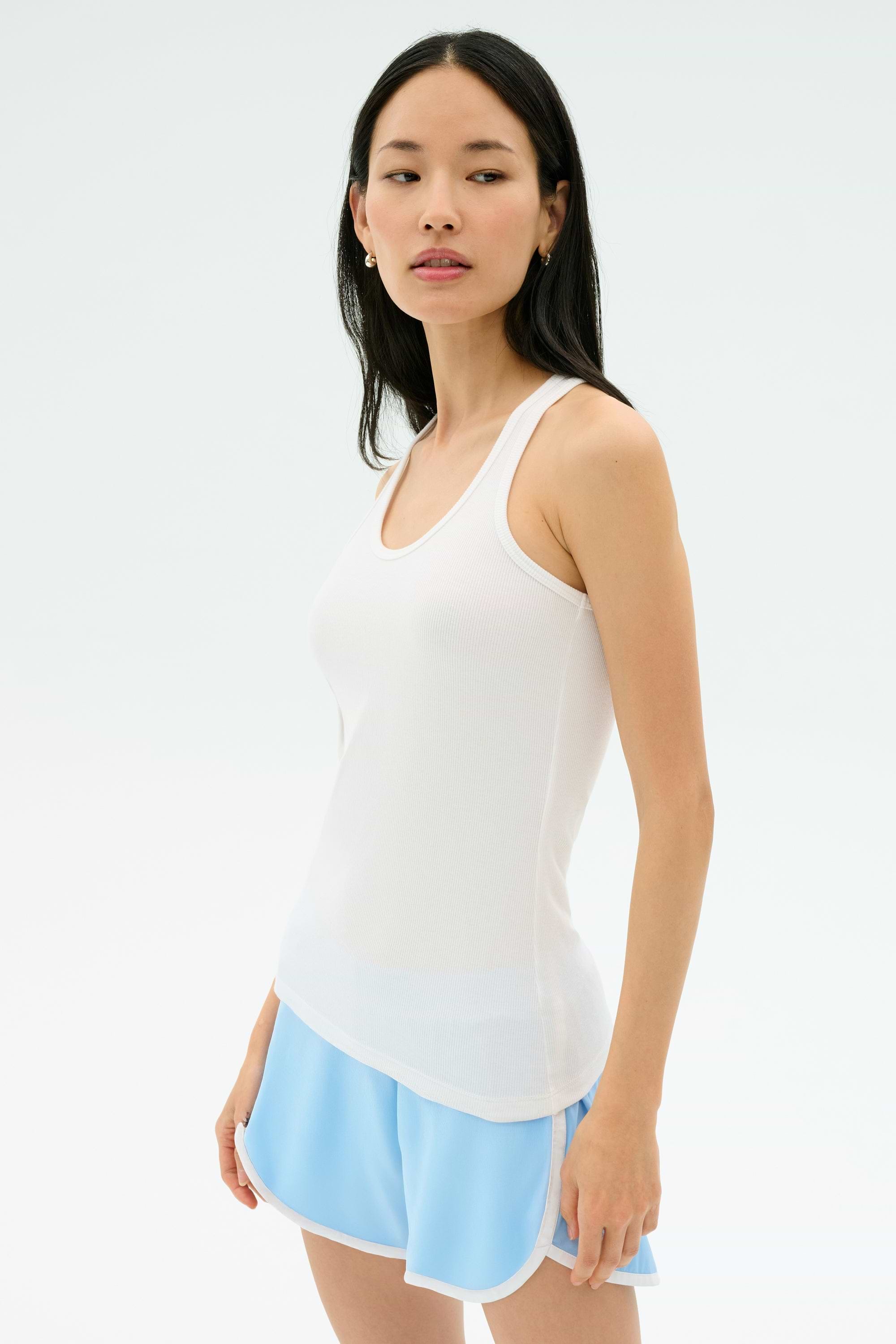 A woman stands against a plain background wearing the Splits59 Ashby Rib Tank in white and light blue athletic shorts, looking slightly to the side.