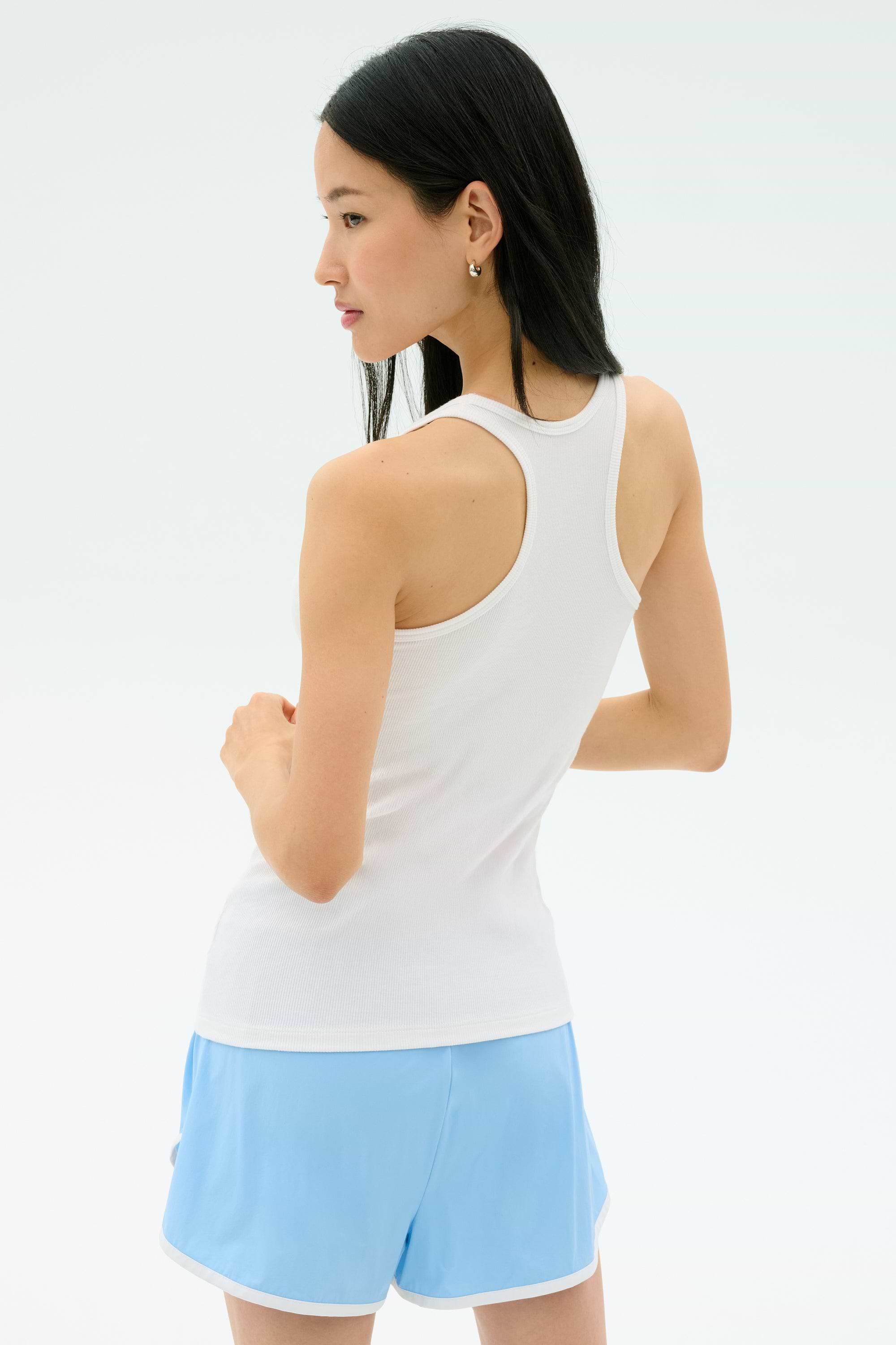A woman with long dark hair models the Splits59 Ashby Rib Tank in white, paired with light blue shorts, standing with her back to the camera against a plain white background.