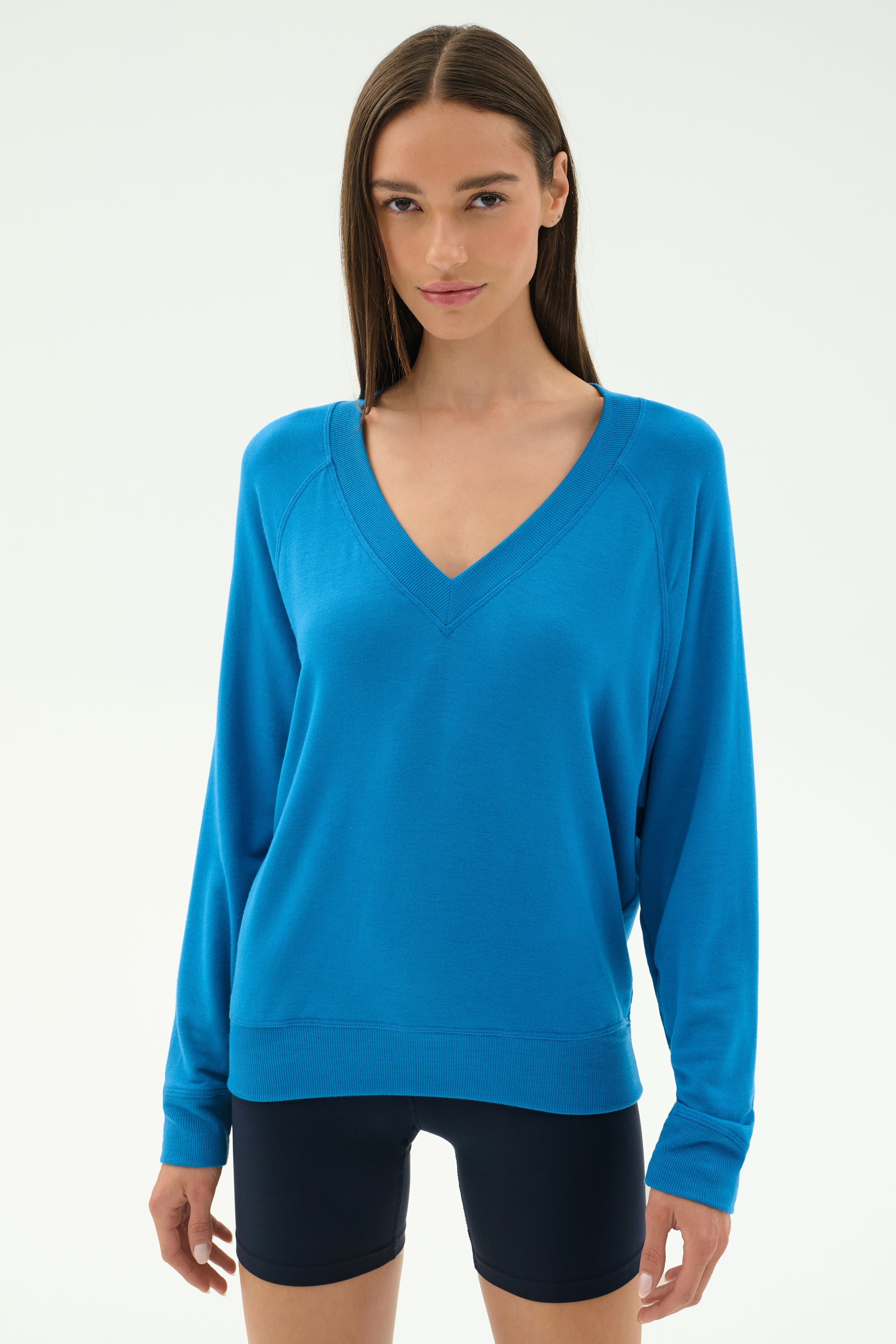 A woman wearing the SPLITS59 Bennie Fleece V Neck Sweatshirt in Stone Blue with black fitted shorts stands against a plain light background.