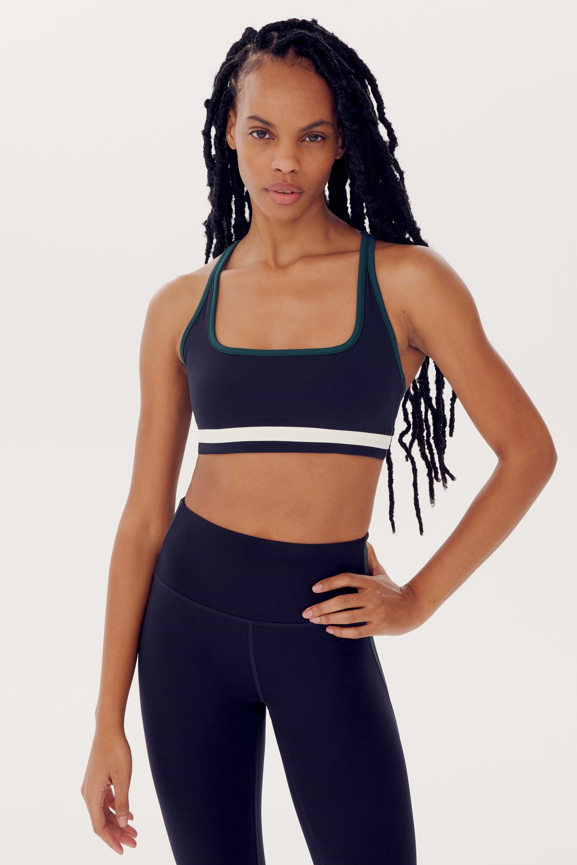 A woman with long braided hair is wearing a Bianca Rigor Bra - Indigo by SPLITS59 and spandex leggings, posing with one hand on her hip against a plain background.
