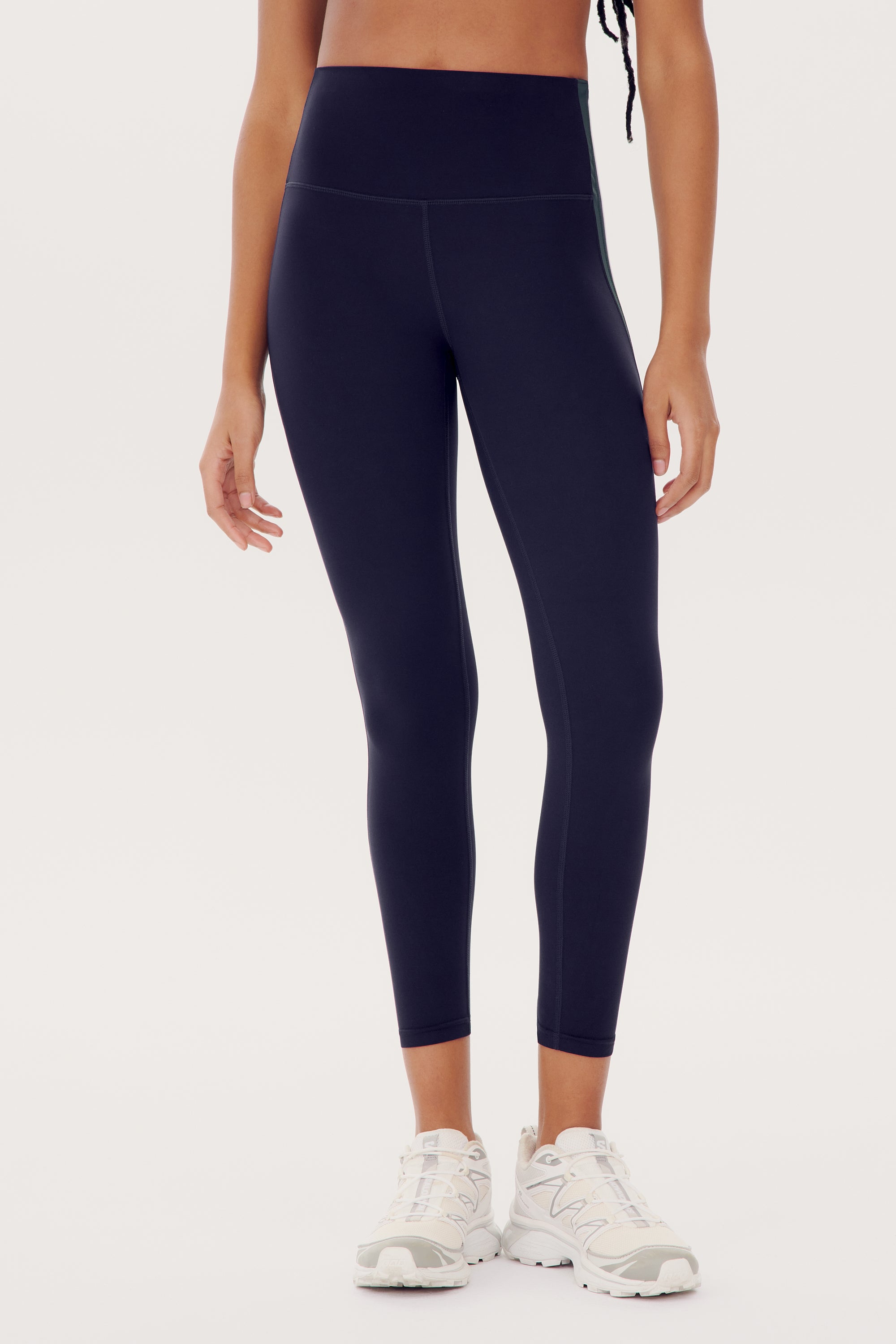 A person is shown from the shoulders down, wearing dark blue Bianca High Waist Rigor 7/8 - Indigo leggings by SPLITS59 and white sneakers against a plain background.