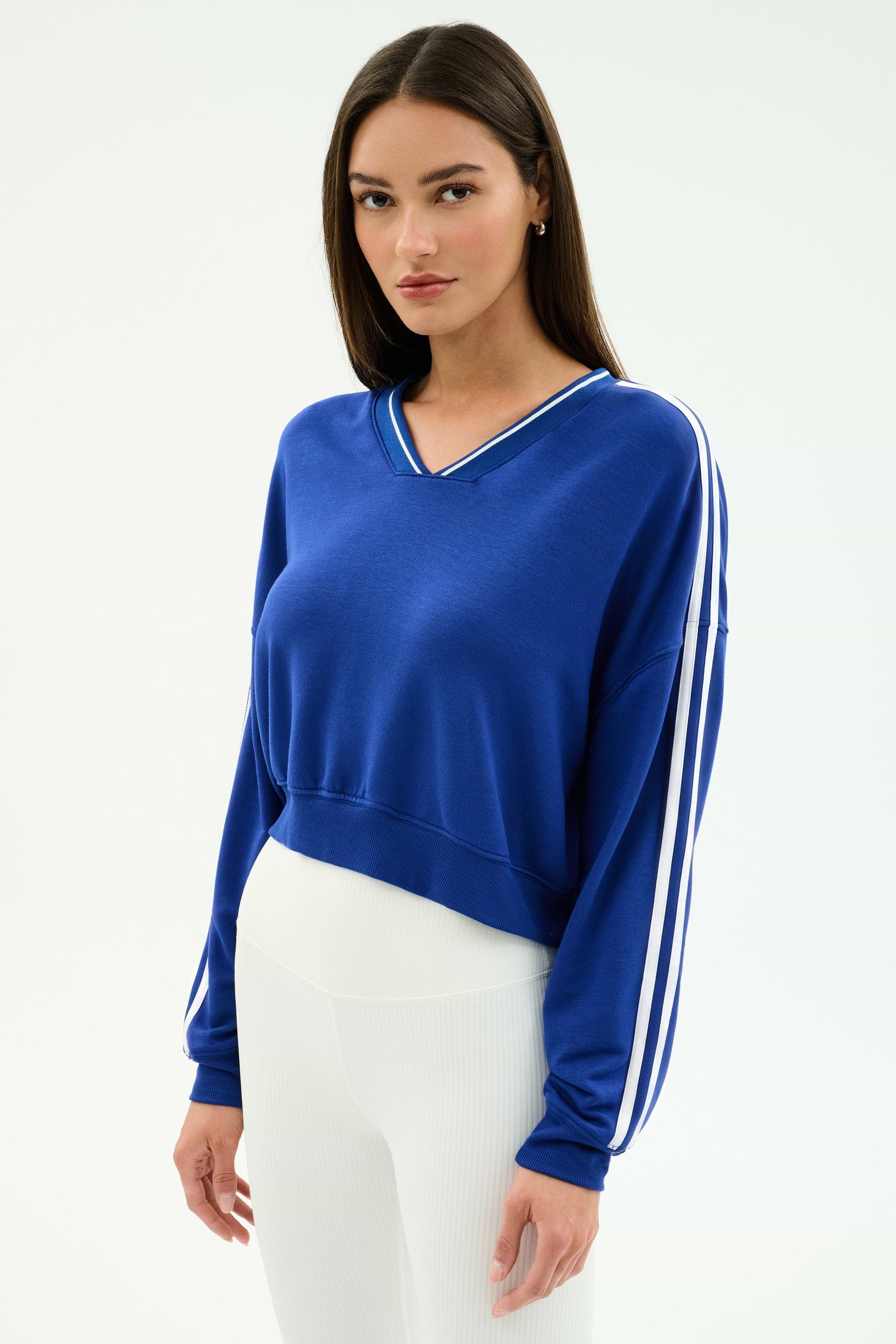 A woman wears the SPLITS59 Ellie Fleece V Neck Sweatshirt in Cobalt/White with white pants, standing against a plain light background for an effortless athletic look.