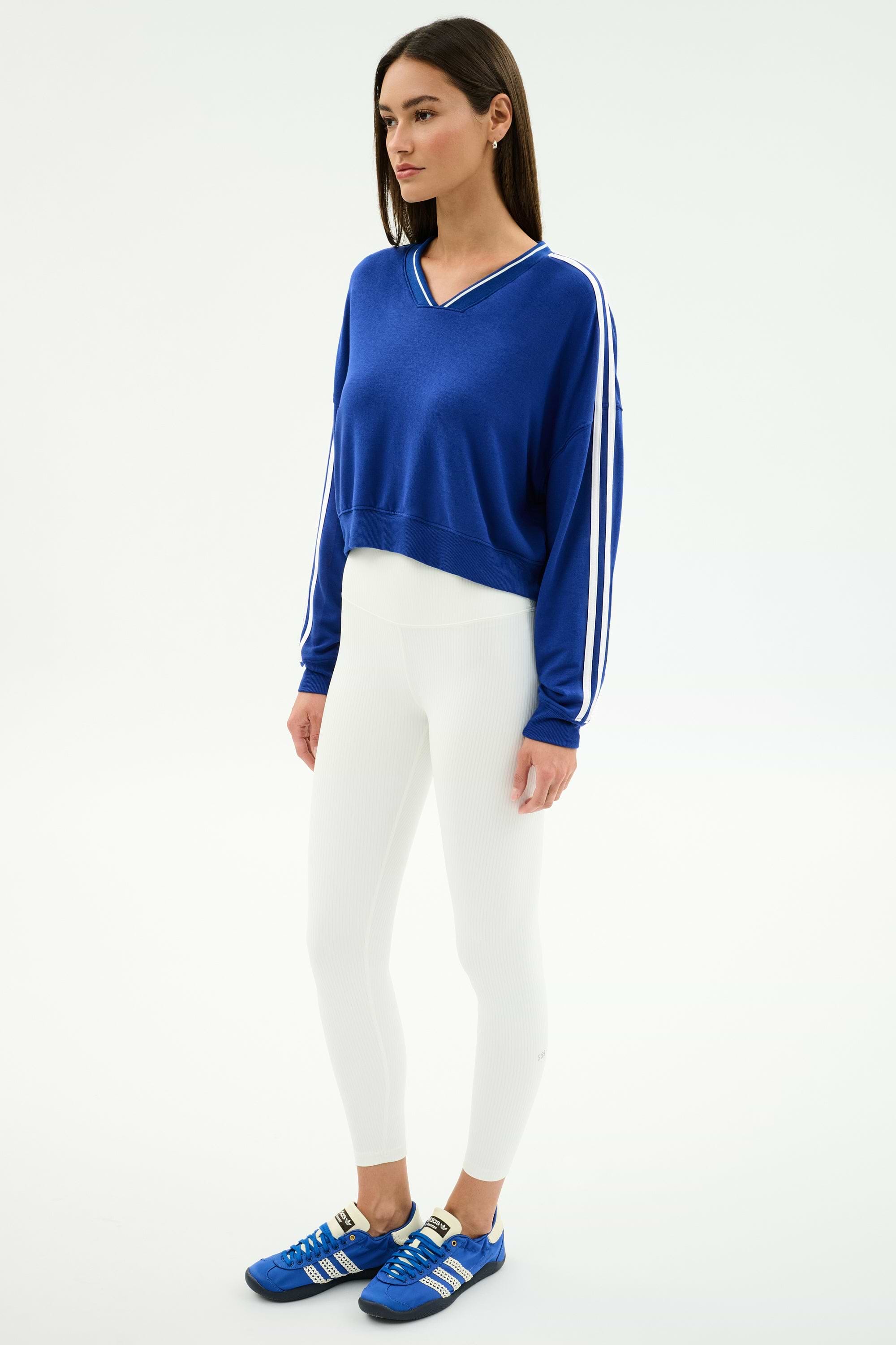 A woman wears the SPLITS59 Ellie Fleece V Neck Sweatshirt in Cobalt/White with white leggings and blue athletic shoes, creating a relaxed athletic look against a plain white background.