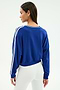 A woman stands with her back to the camera, wearing the SPLITS59 Ellie Fleece V Neck Sweatshirt in cobalt and white, paired with white high-waisted pants for an athletic look.