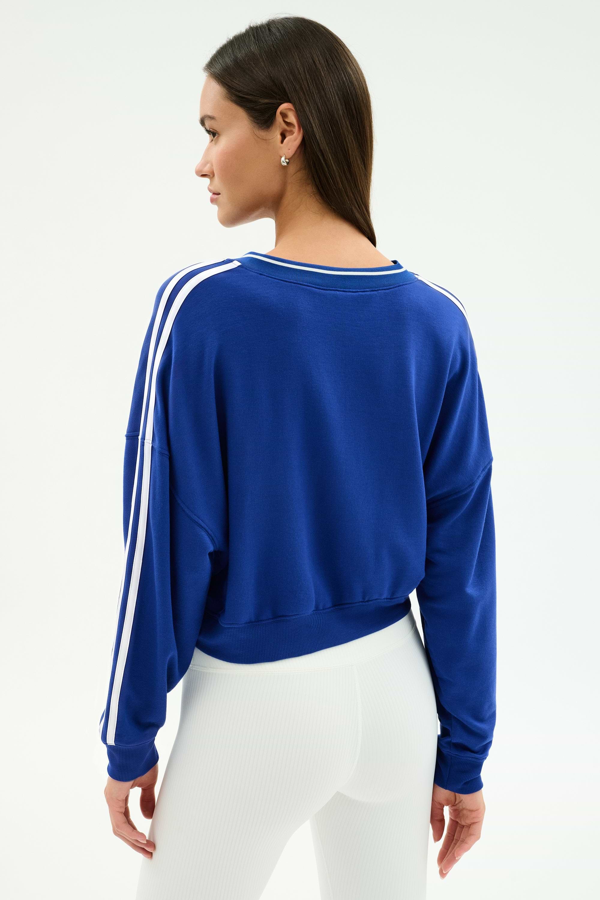 A woman stands with her back to the camera, wearing the SPLITS59 Ellie Fleece V Neck Sweatshirt in cobalt and white, paired with white high-waisted pants for an athletic look.