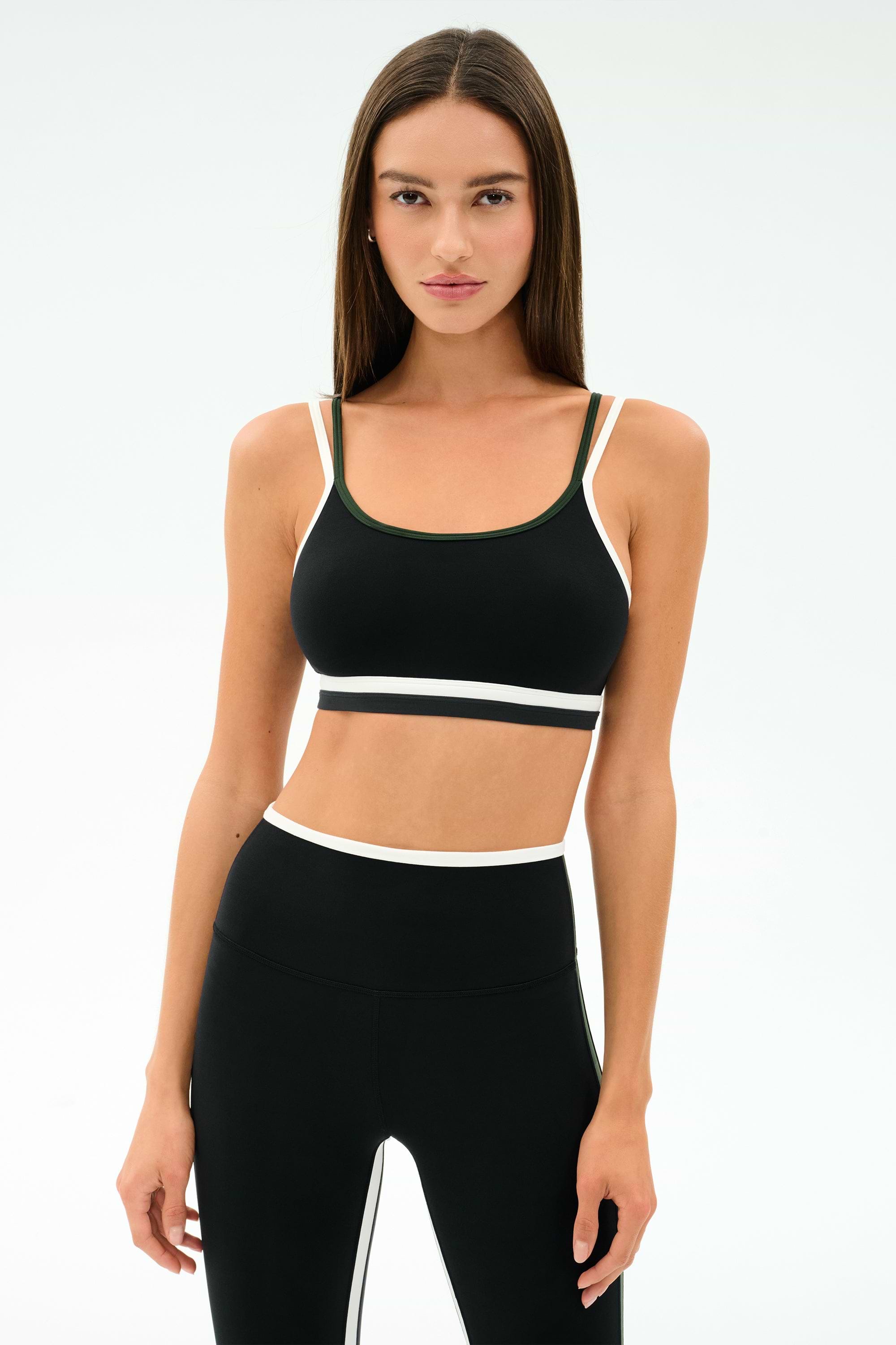 A woman stands against a plain background, wearing the SPLITS59 Eva Rigor Double Strap Bra in black with matching high-waisted leggings with white trim, looking directly at the camera.