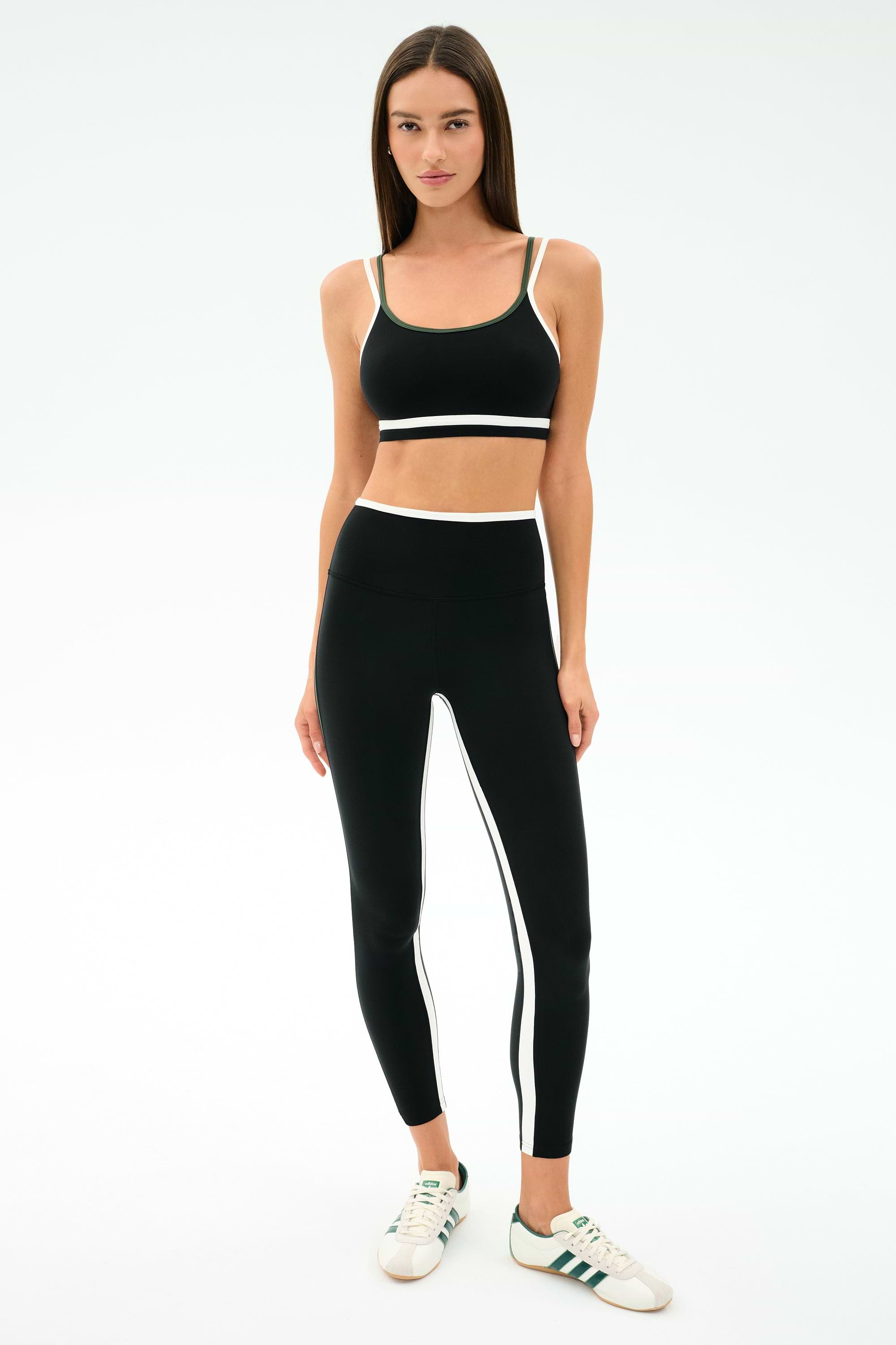 A woman faces forward wearing the SPLITS59 Eva Rigor Double Strap Bra in Black/Military, paired with matching black leggings featuring white stripes, and white sneakers with green accents.