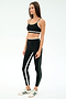 A woman stands in a studio wearing the SPLITS59 Eva Rigor Double Strap Bra in Black/Military, paired with matching black leggings featuring white and green stripes and white sneakers.