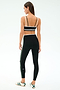 A woman stands with her back to the camera, wearing the SPLITS59 Eva Rigor Double Strap Bra in Black/Military, matching black leggings with side stripes, and white sneakers featuring green accents.