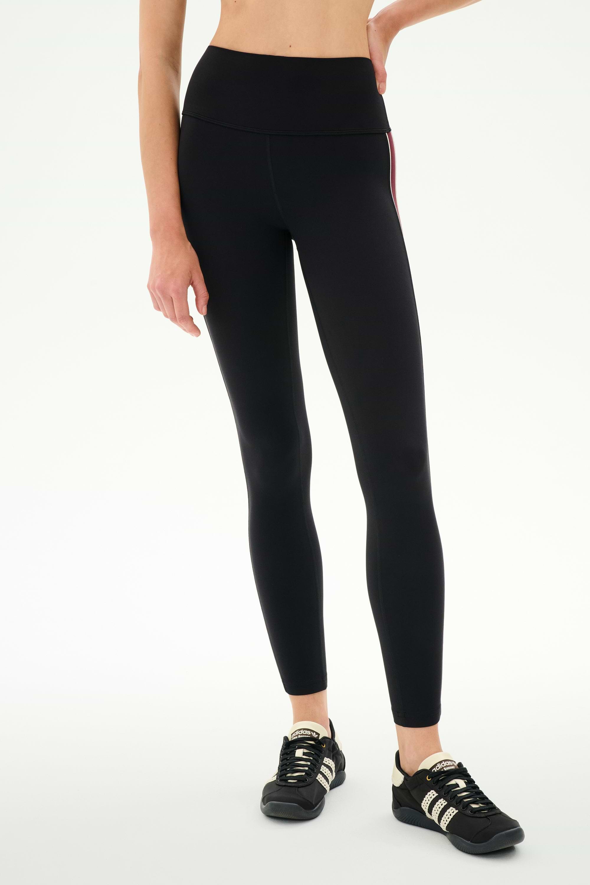 A person wearing SPLITS59 Margot Rigor Legging in Black/Dark Cherry and black athletic shoes stands against a plain white background, with one arm resting on their hip.