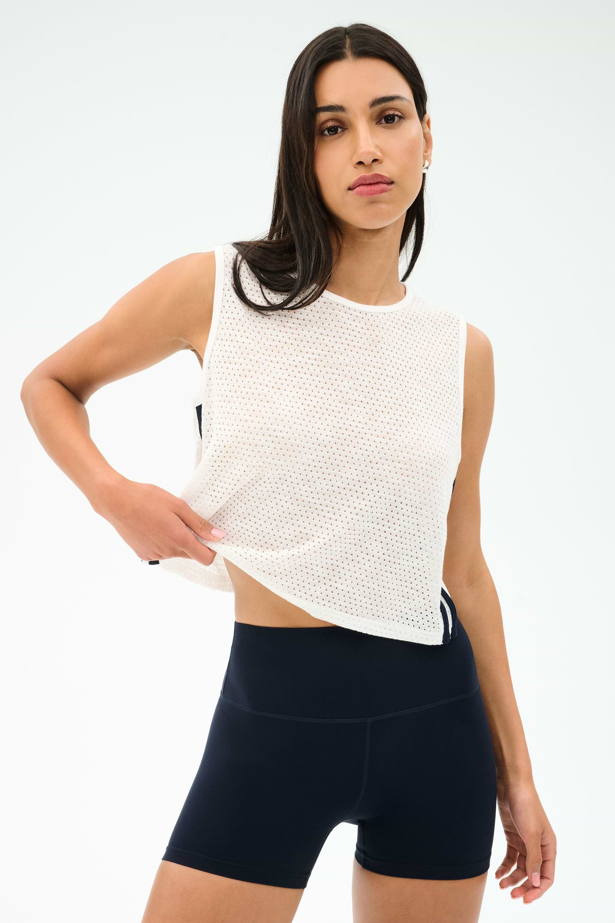A woman wears the SPLITS59 Logan Mesh Tank With Stripe in White/Indigo and black athletic shorts, posing against a light background—an ideal breathable sportswear choice for your next workout.