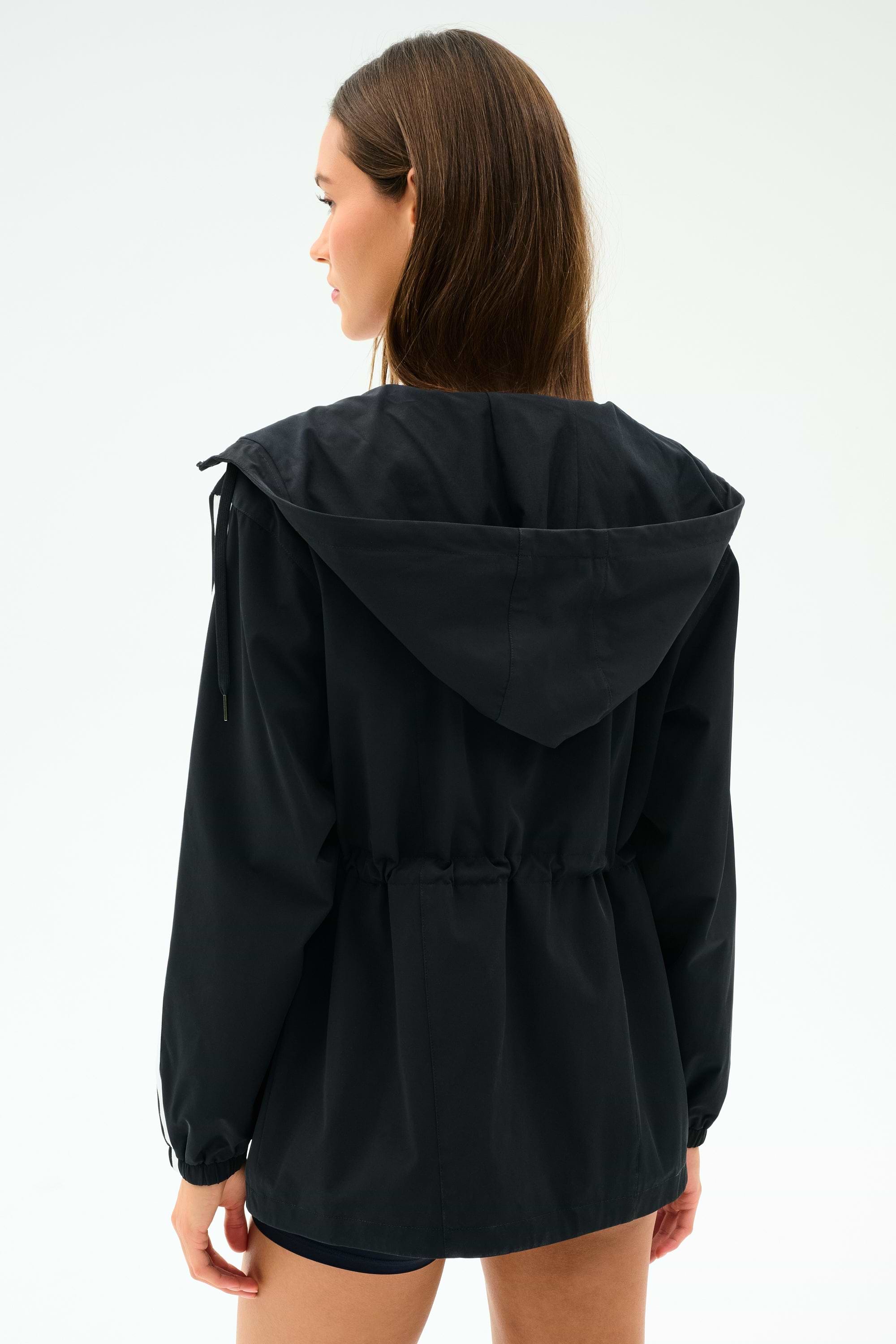 A woman faces away, wearing the SPLITS59 Reggie Water Repellent Jacket in black/white, featuring a cinched waist and sleek nylon fabric.