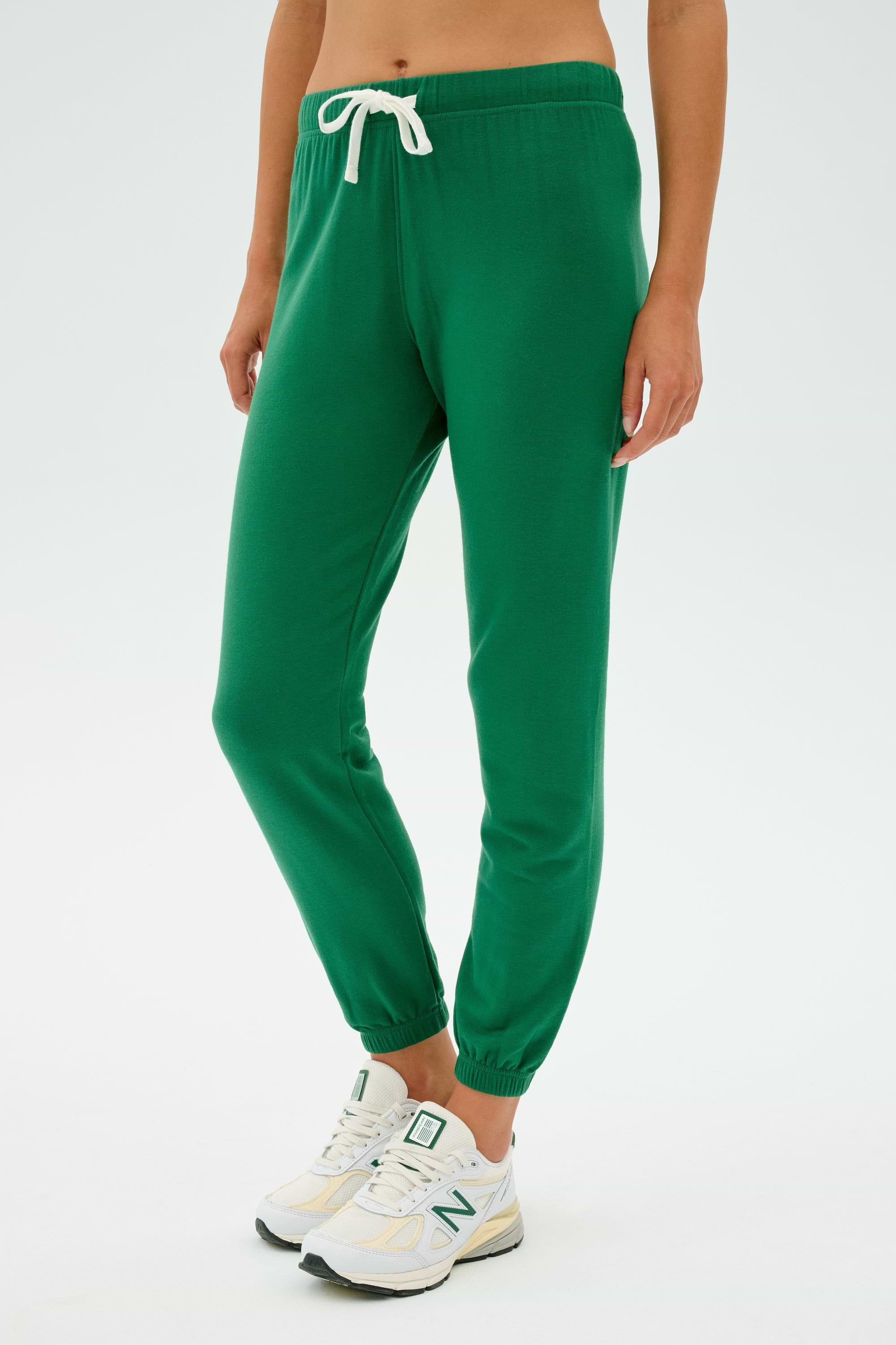 A person dressed in SPLITS59's Sonja Fleece Sweatpant in Arugula and white sneakers stands against a plain background.
