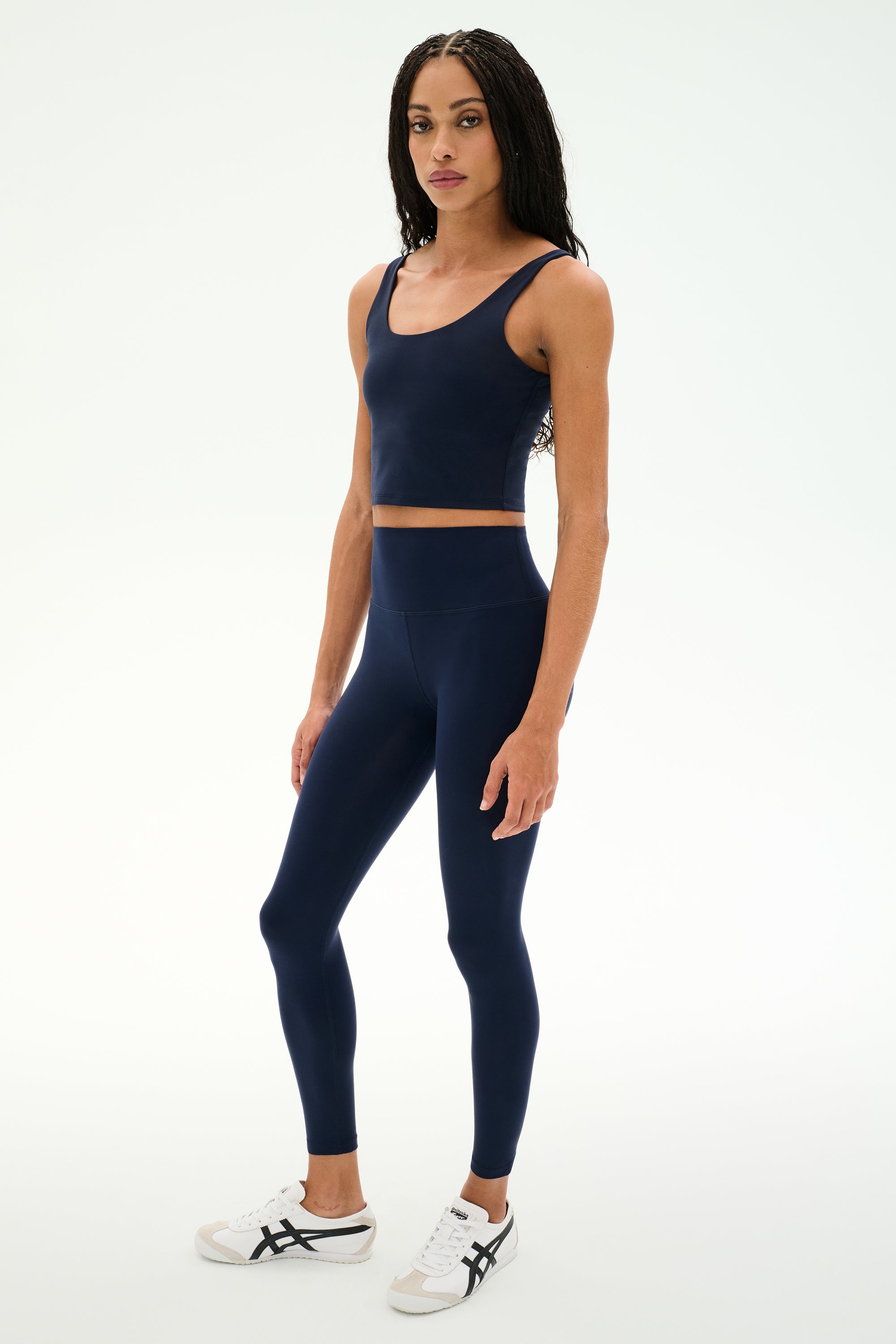 A person is wearing the SPLITS59 Sprint High Waist Rigor 7/8 leggings in Indigo, crafted from Rigor fabric and paired with a matching navy blue sports crop top and white sneakers, standing against a plain white background.