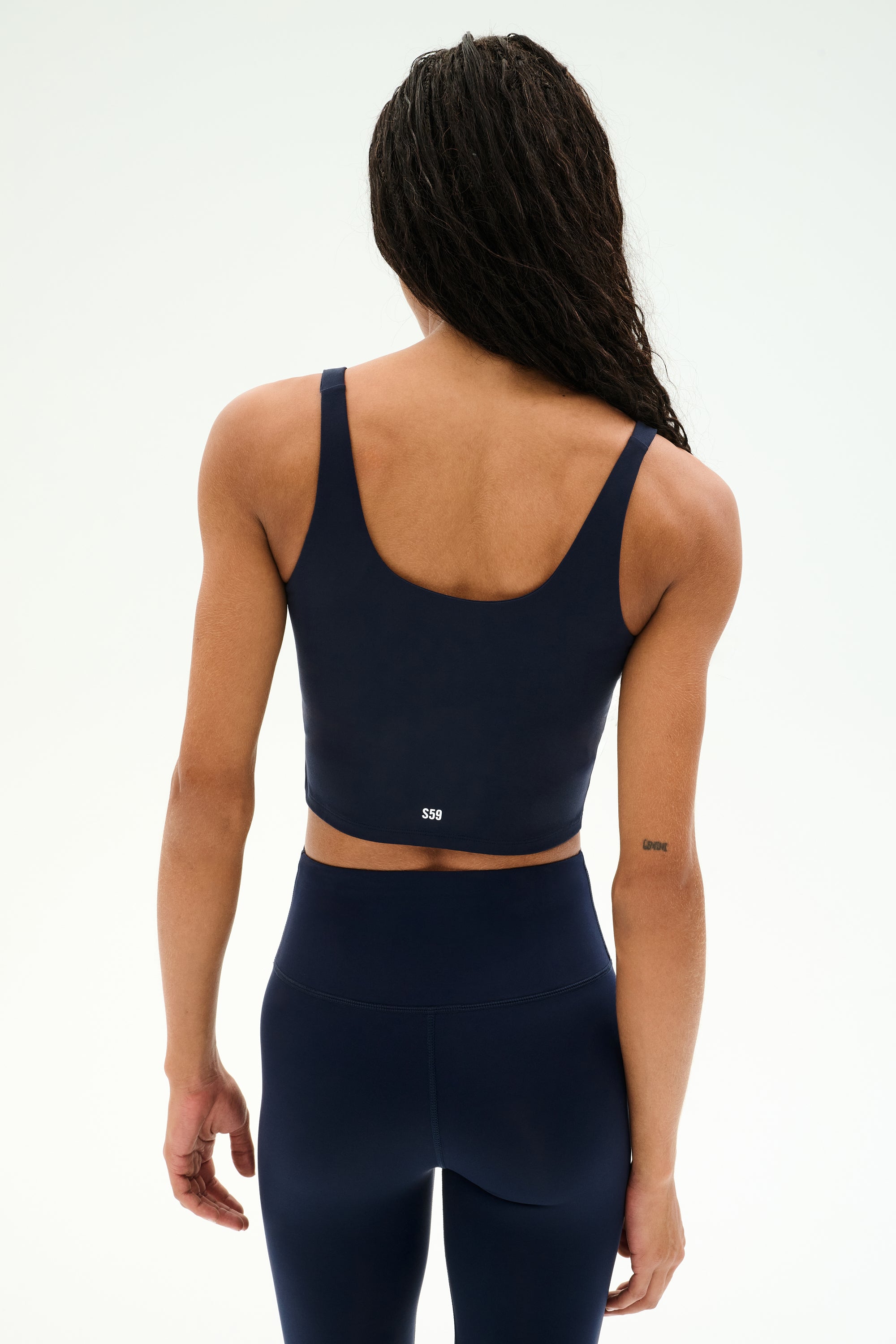 A woman with long braided hair, seen from the back, wears the SPLITS59 Sprint Rigor Bralette in Indigo—crafted for multi-sport performance—paired with matching high-waisted leggings against a plain white background.