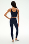 A person is shown from the back, standing on a white background, wearing SPLITS59's Sprint High Waist Rigor 7/8 leggings in Indigo—a navy blue shade—paired with a matching athletic crop top and white athletic shoes.