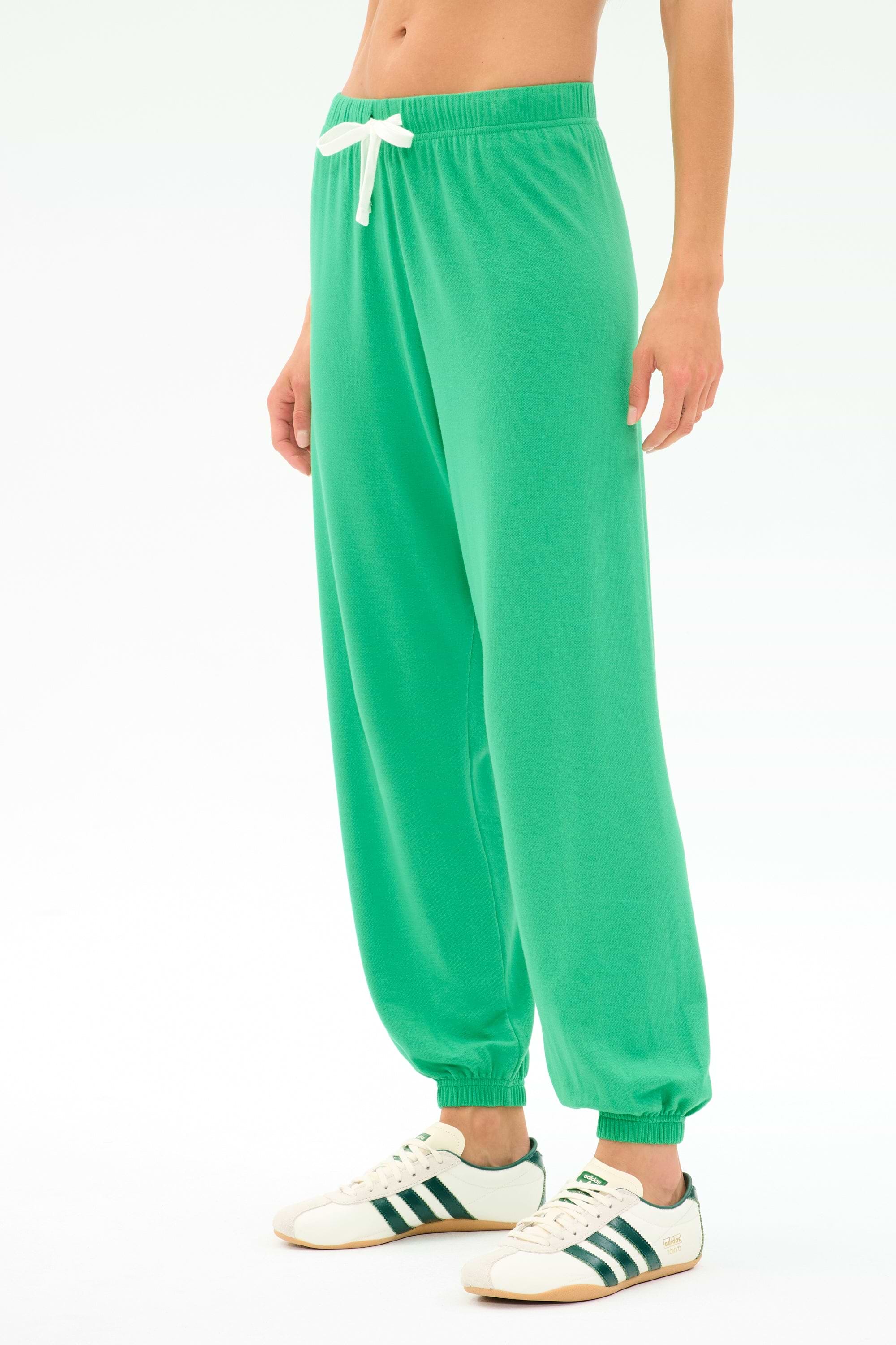 A person wearing SPLITS59’s Andie Oversized Fleece Sweatpant in Spearmint with a white drawstring and white sneakers with green stripes stands against a plain white background.