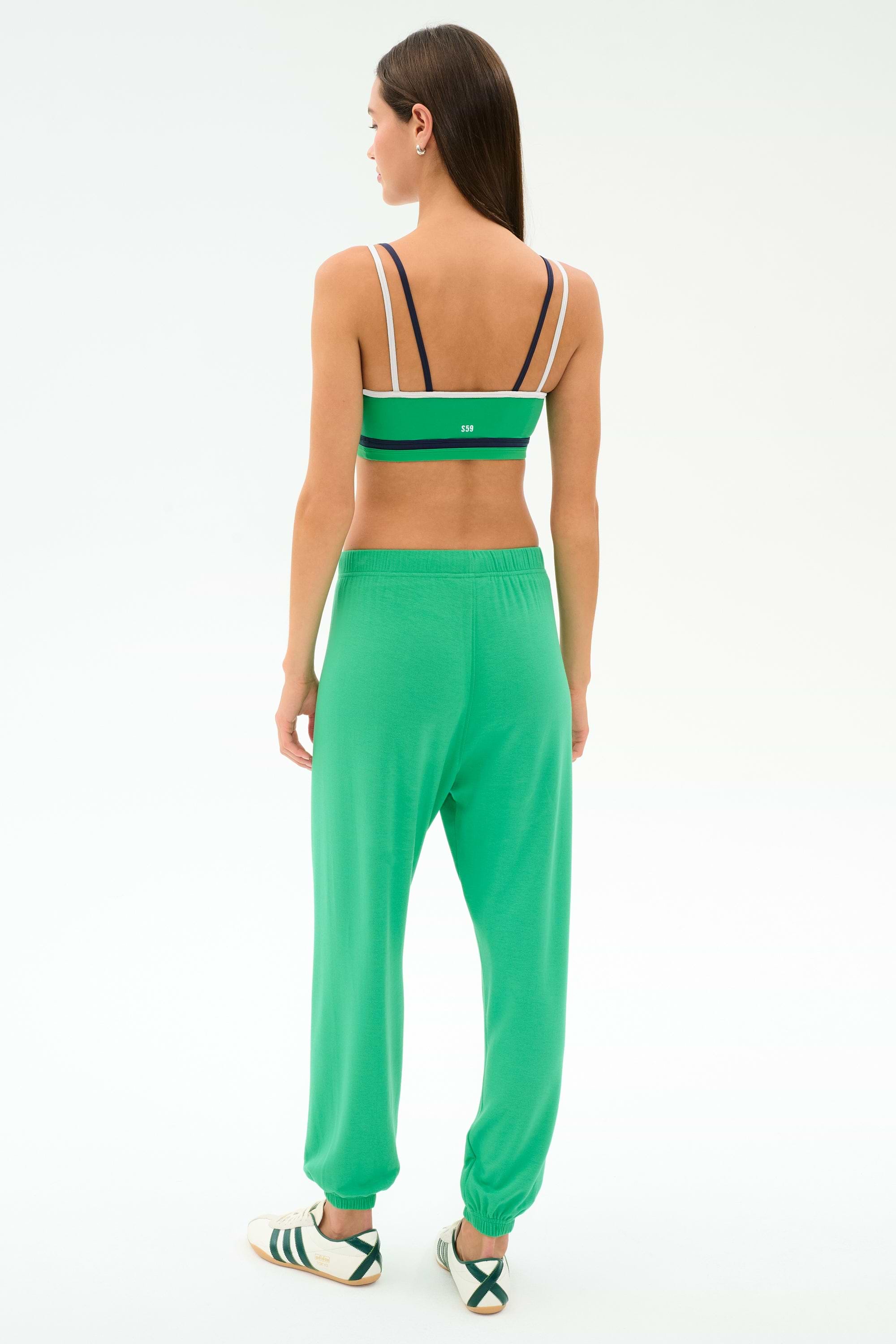 A woman faces away, wearing SPLITS59’s Andie Oversized Fleece Sweatpant in spearmint with a green sports bra and white sneakers with black stripes, all set against a plain white background.