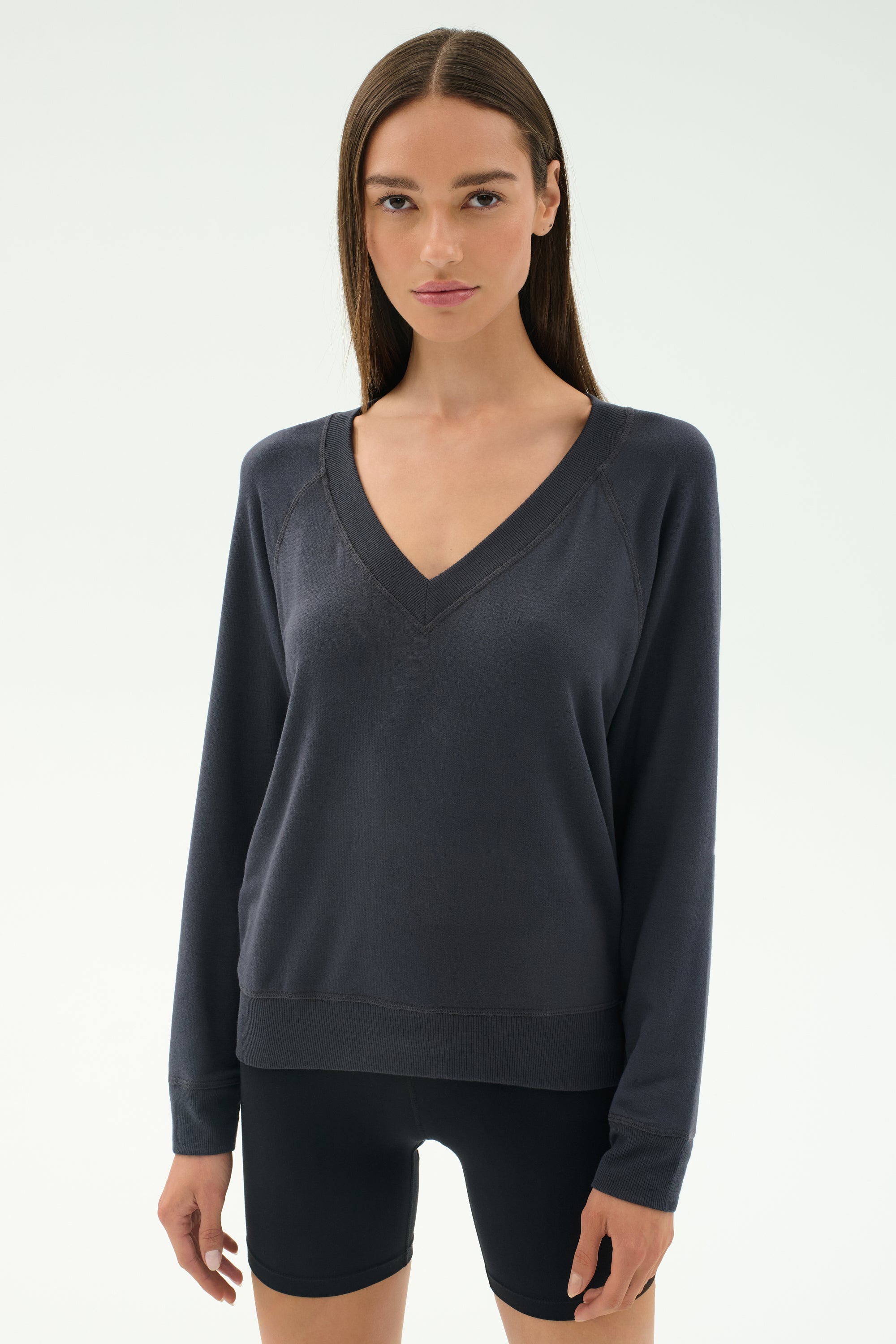 A woman stands against a light background, wearing the SPLITS59 Bennie Fleece V Neck Sweatshirt in Graphite and black shorts. This chic pullover is crafted in Los Angeles.
