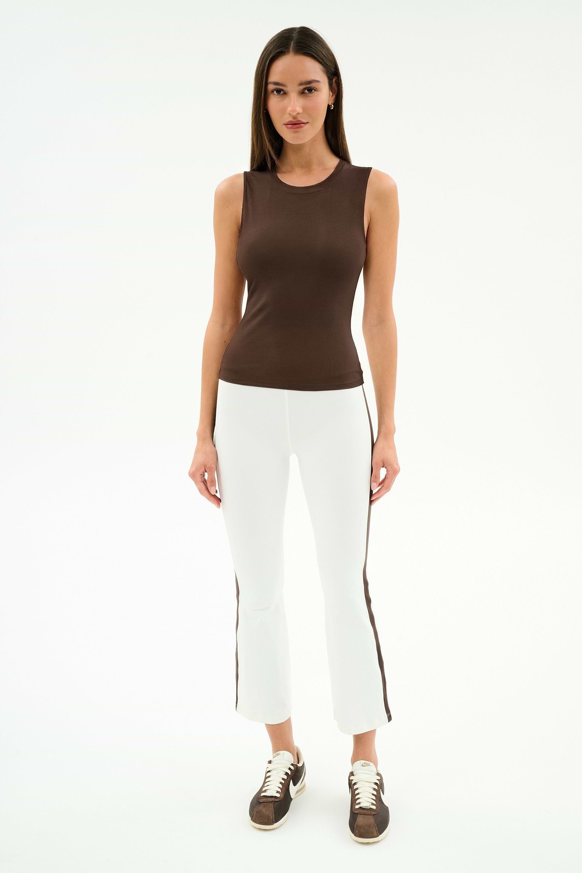 A woman stands against a white background wearing a sleeveless brown top, brown sneakers, and SPLITS59 Raquel High Waist Supplex Crop leggings in White/Dark Chocolate.