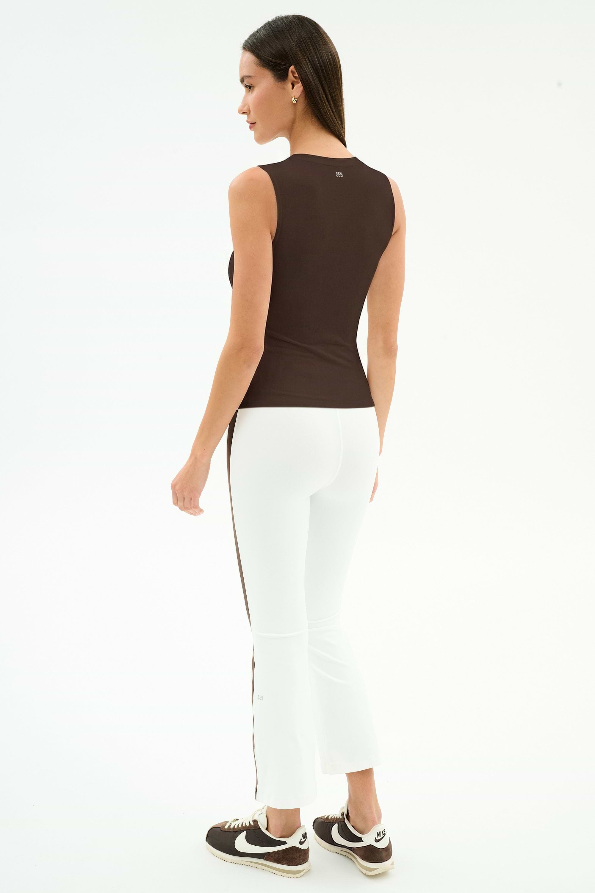 A woman faces away on a plain white background, wearing the SPLITS59 Carter Airweight Lite Tank in Dark Chocolate, paired with white pants featuring a brown stripe and brown Nike sneakers.