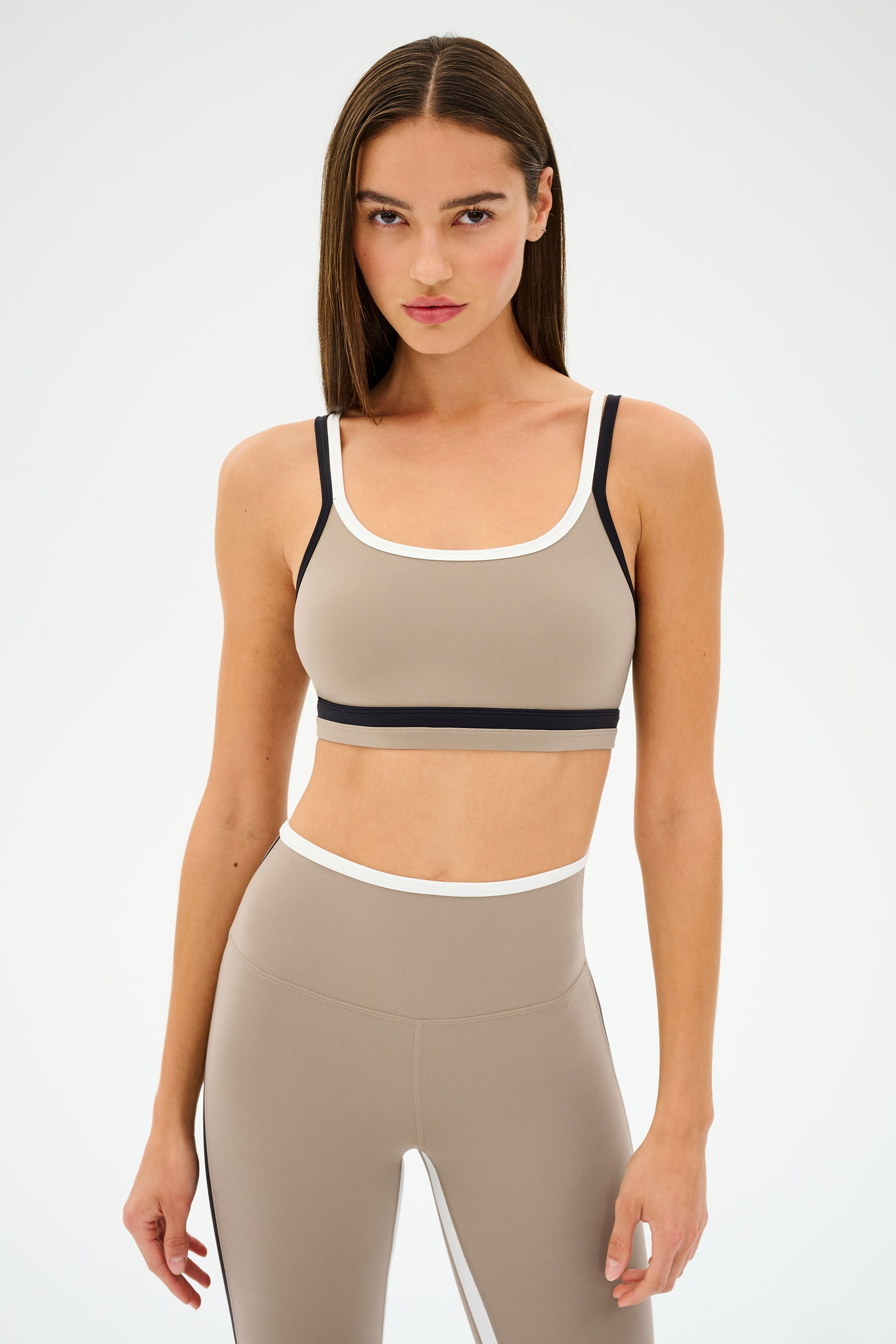 A model in a beige and black athletic outfit showcases the SPLITS59 Eva Rigor Double Strap Bra in Latte/Black against a plain backdrop.