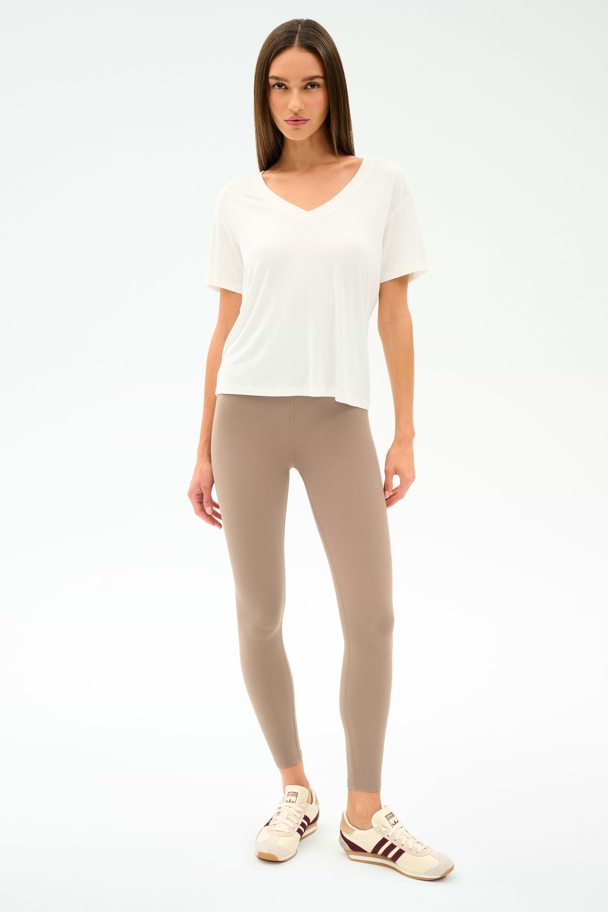 A woman faces forward against a plain white background, wearing the SPLITS59 Georgia Jersey V-Neck in white, styled with beige leggings and beige sneakers with maroon stripes.