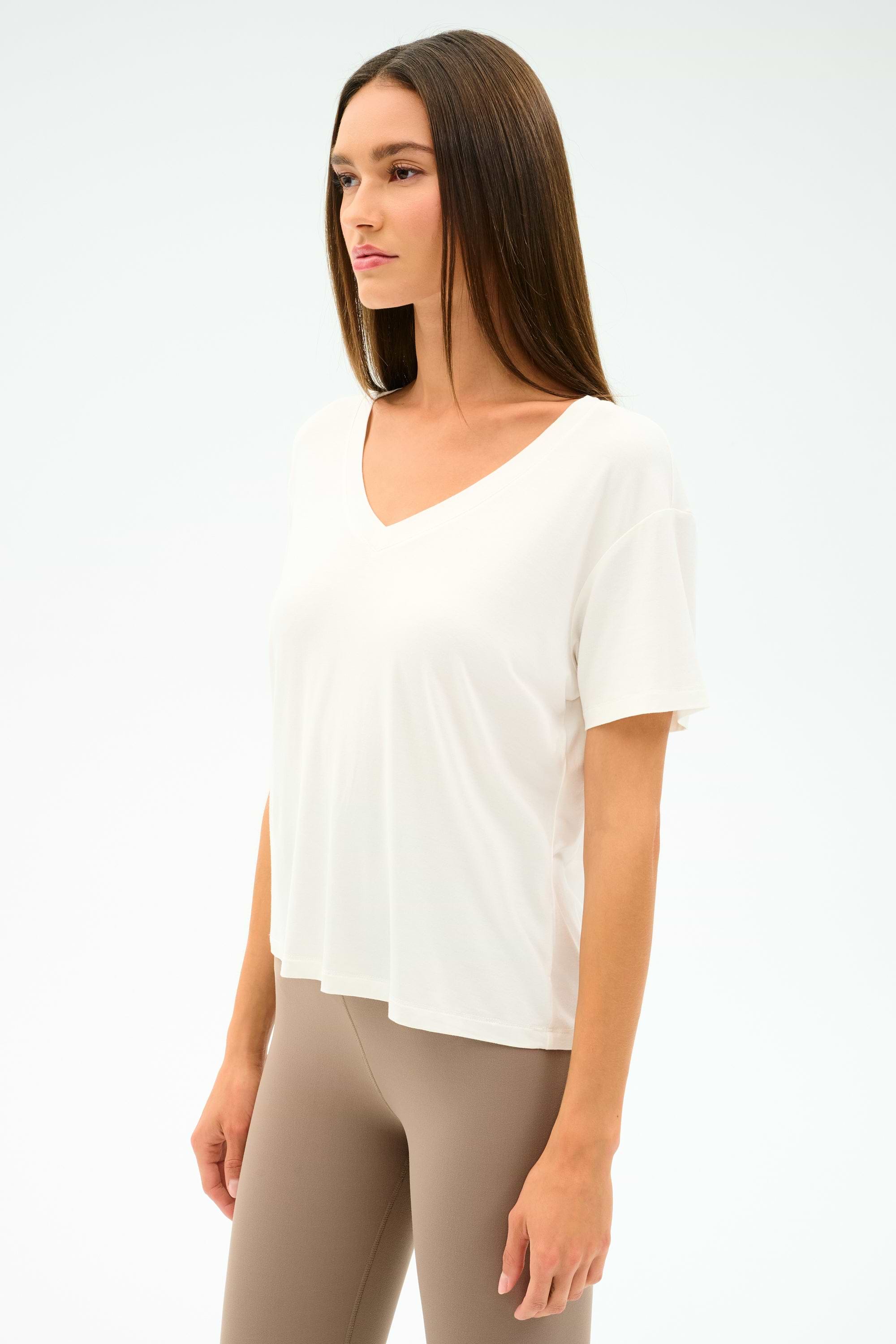 A woman stands against a plain light background wearing the SPLITS59 Georgia Jersey V-Neck in white, a loose-fitting T-shirt crafted from a soft viscose blend, paired with beige leggings.