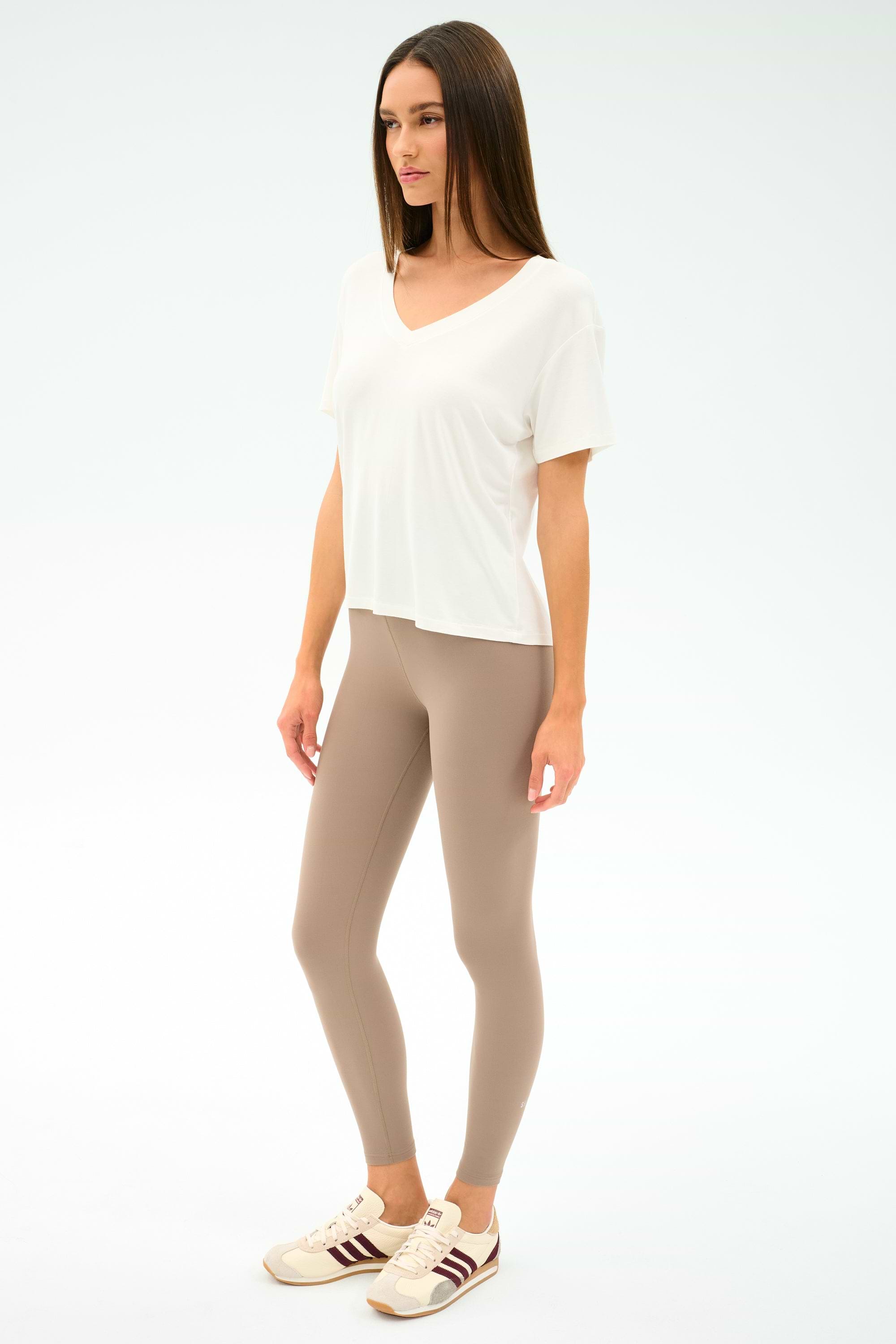 A woman stands against a plain background wearing the SPLITS59 Georgia Jersey V-Neck in white, paired with beige leggings and beige sneakers featuring brown stripes.