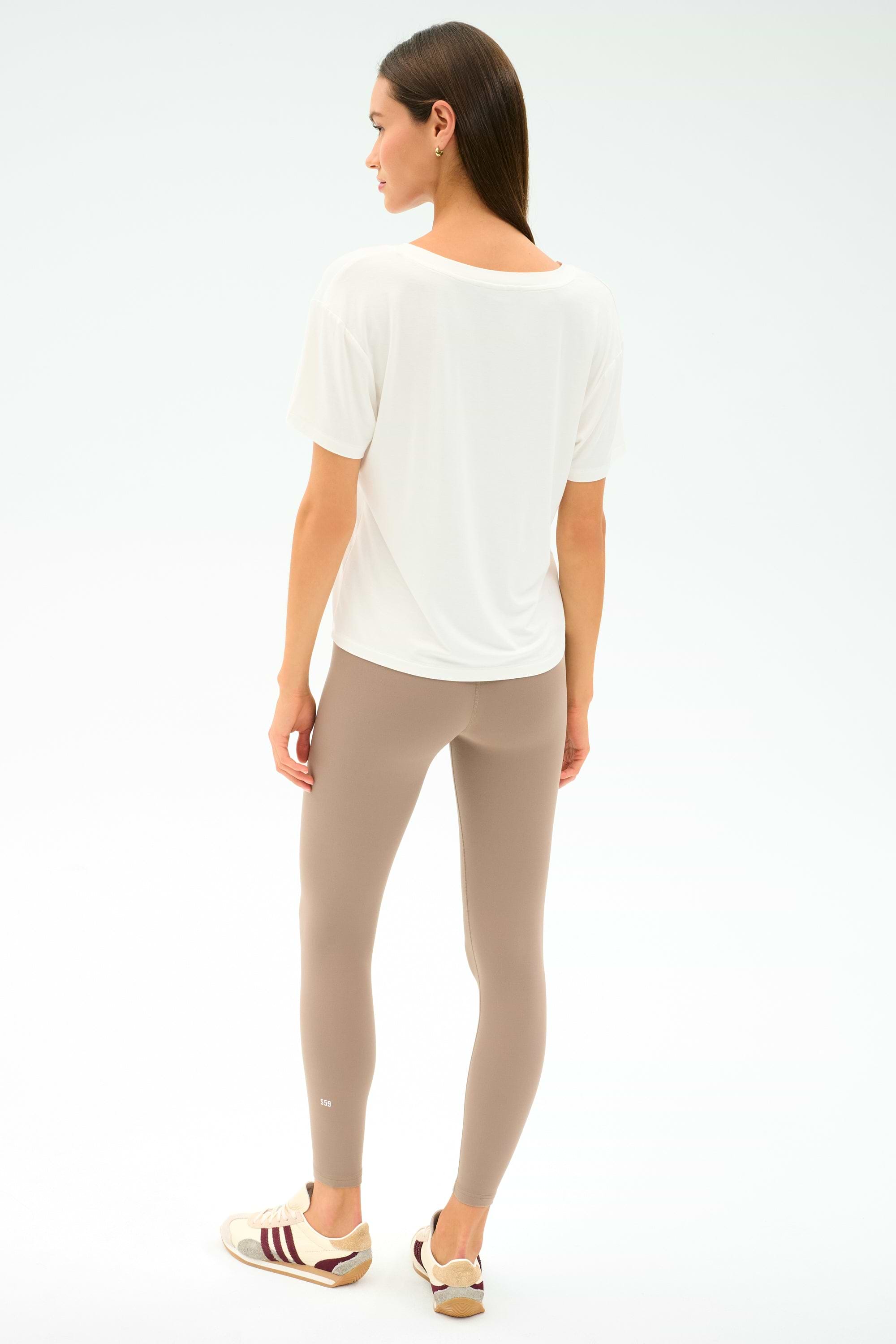 A woman faces away, wearing the SPLITS59 Georgia Jersey V-Neck in white and taupe spandex-blend leggings, paired with beige sneakers featuring maroon stripes, set against a plain light background.